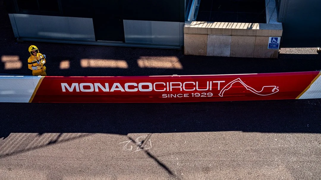 Red banner with white text reading "Monaco Circuit Since 1929", with a graphic outline of a race track, placed outside a building. A person in a yellow safety suit and helmet is standing nearby.