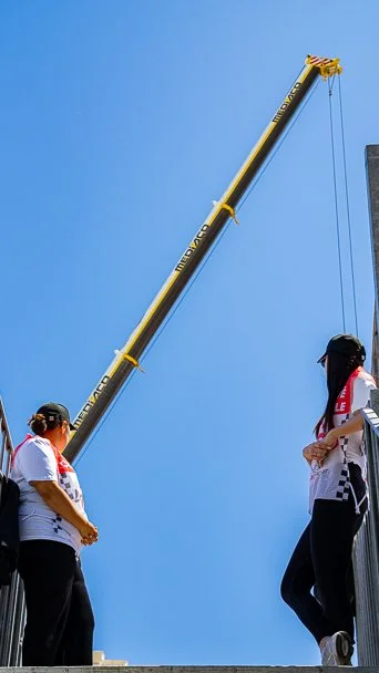 Two women standing near a tall yellow construction crane against a clear blue sky.
