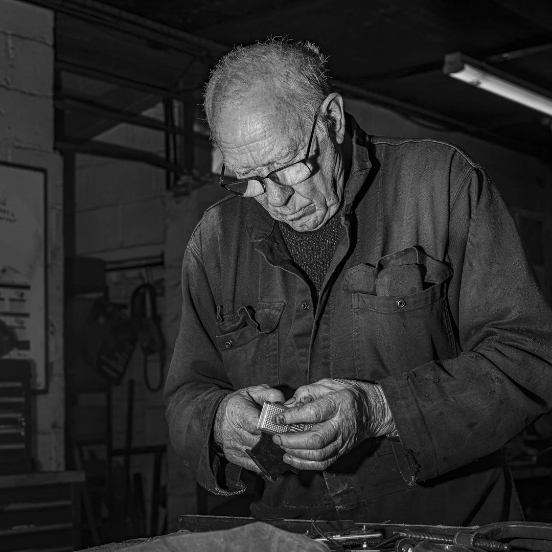 An elderly man with glasses welding metal in a workshop.