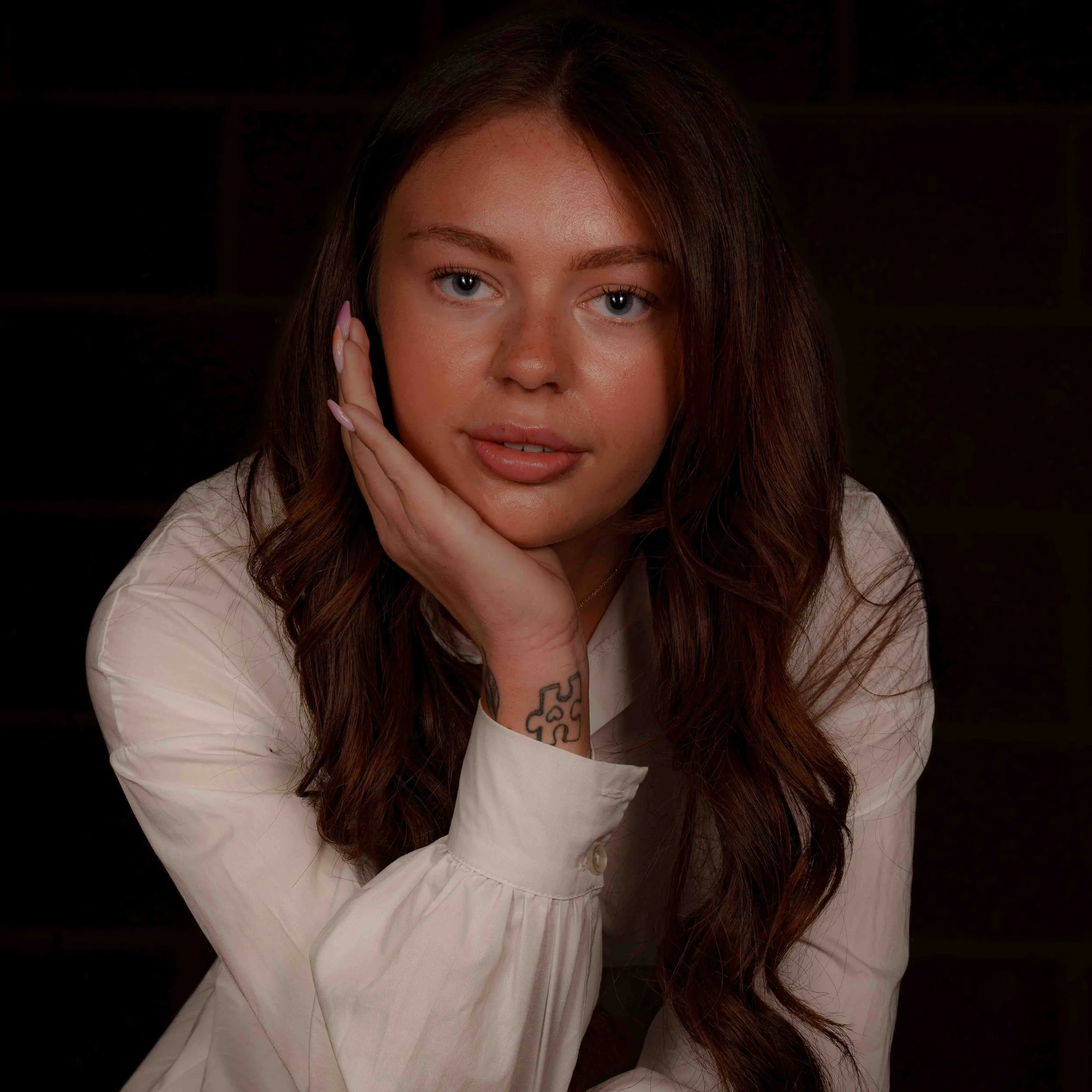 A woman with long brown hair, blue eyes, and freckles resting her chin on her hand, wearing a white shirt against a dark background.