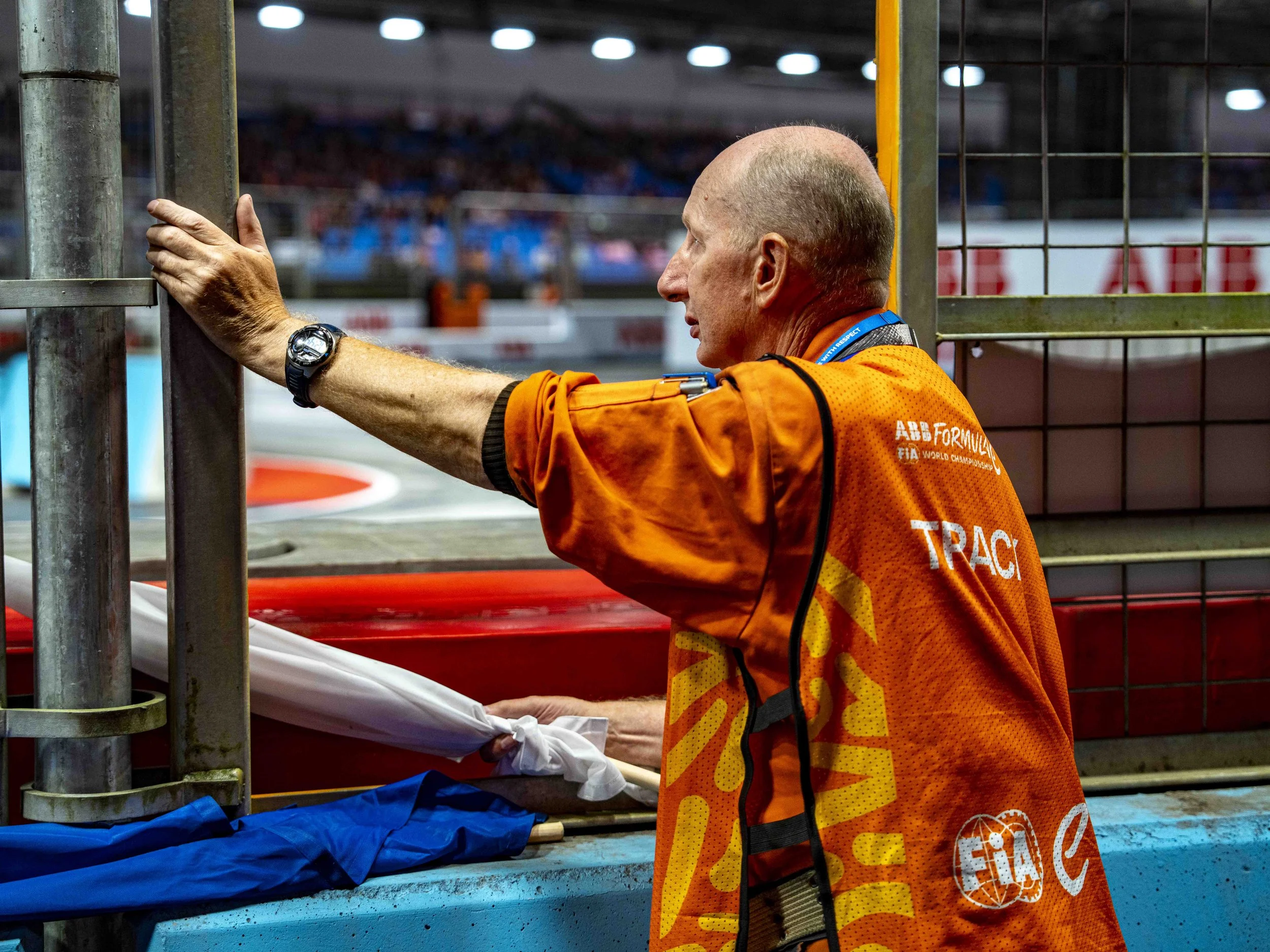 A man wearing an orange FIA Formula E jacket and a wristwatch is standing at a racetrack, holding onto a metal railing and looking at the track.