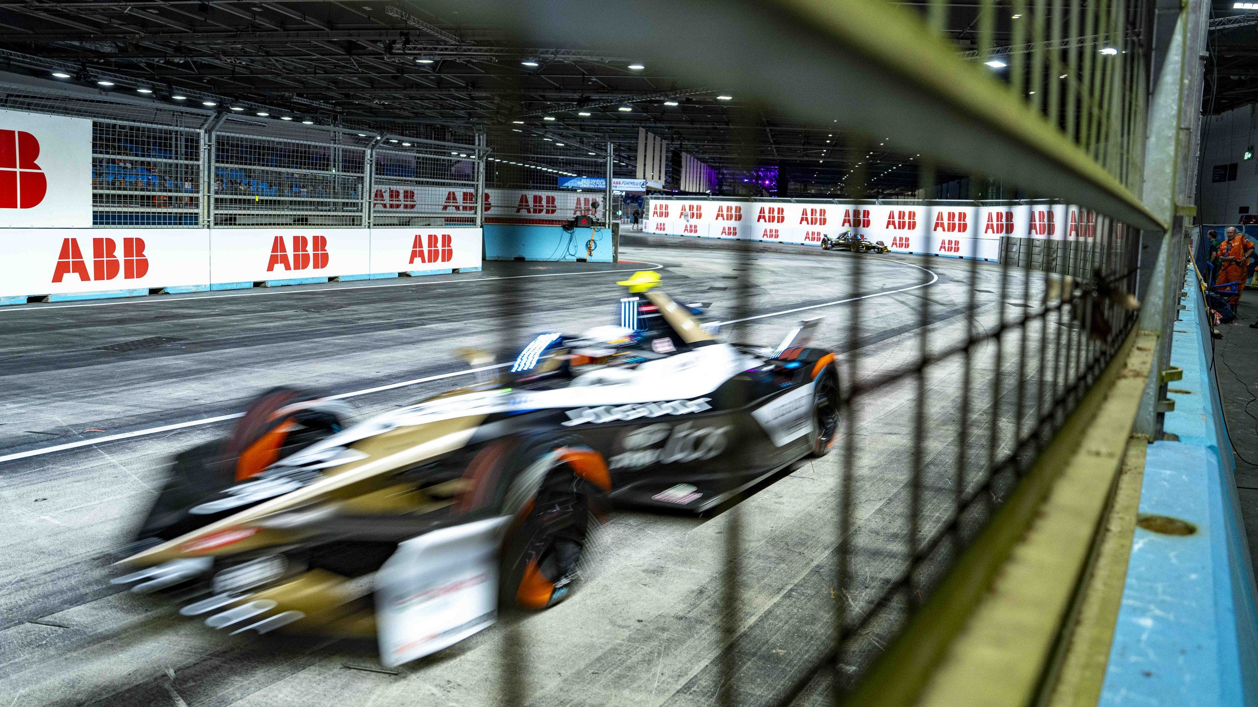 A Formula E race car speeding through a pit lane with motion blur, seen through a metal fence with a blue and black wall on the side, and illuminated by bright overhead lights.