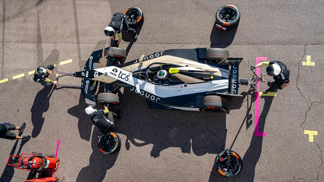 Overhead view of a Jaguar racing car in the pit with four crew members working on it and three additional tires.