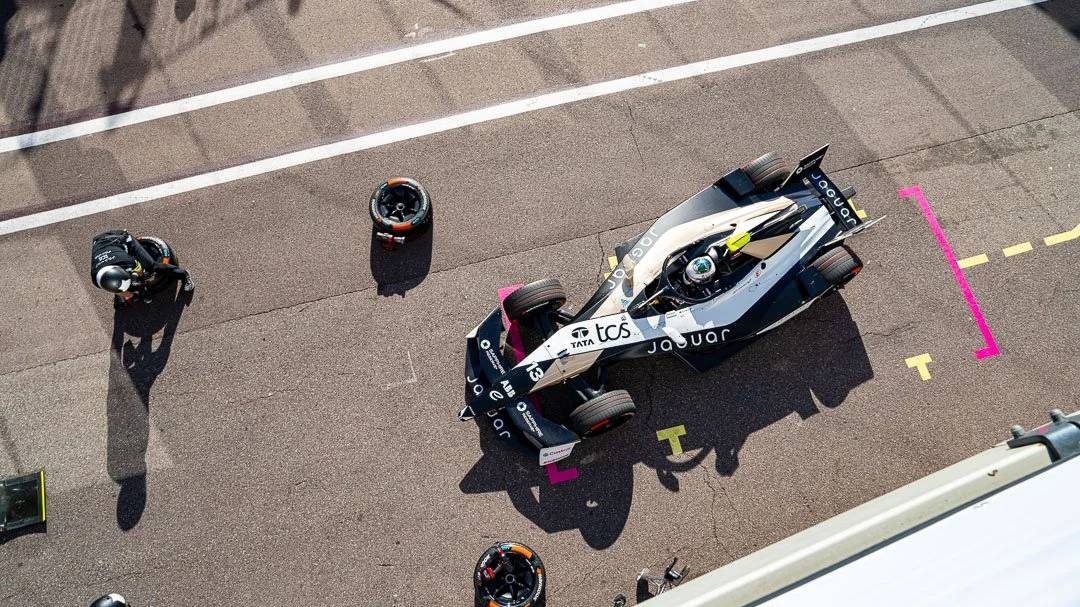 Top-down view of a Formula E race car in the pit lane, with crew members and tires nearby.