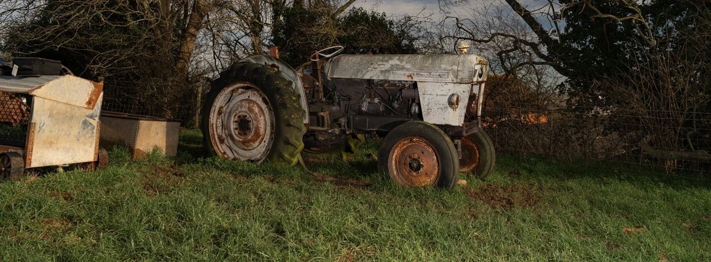 Old, rusty tractor on a grassy field with trees and a fence in the background.