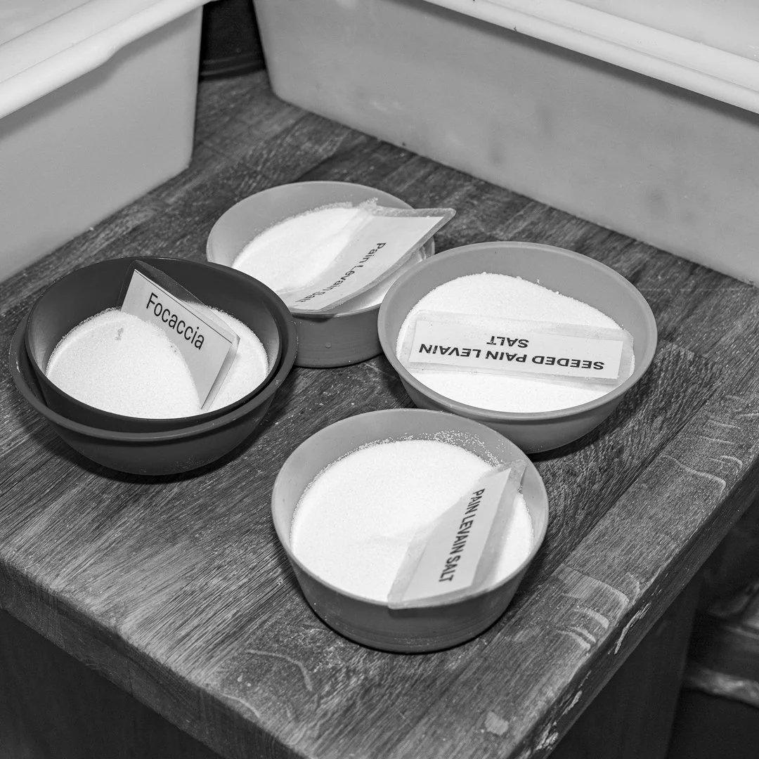 Four bowls with various seasonings and labels, including fennel, ground cumin, seasoned plain salt, and pink lemon salt, on a wooden surface.