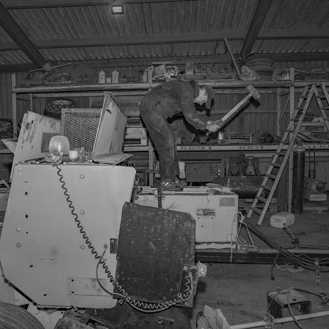 A person working in a cluttered workshop with tools, ladders, and equipment, using a sledgehammer.