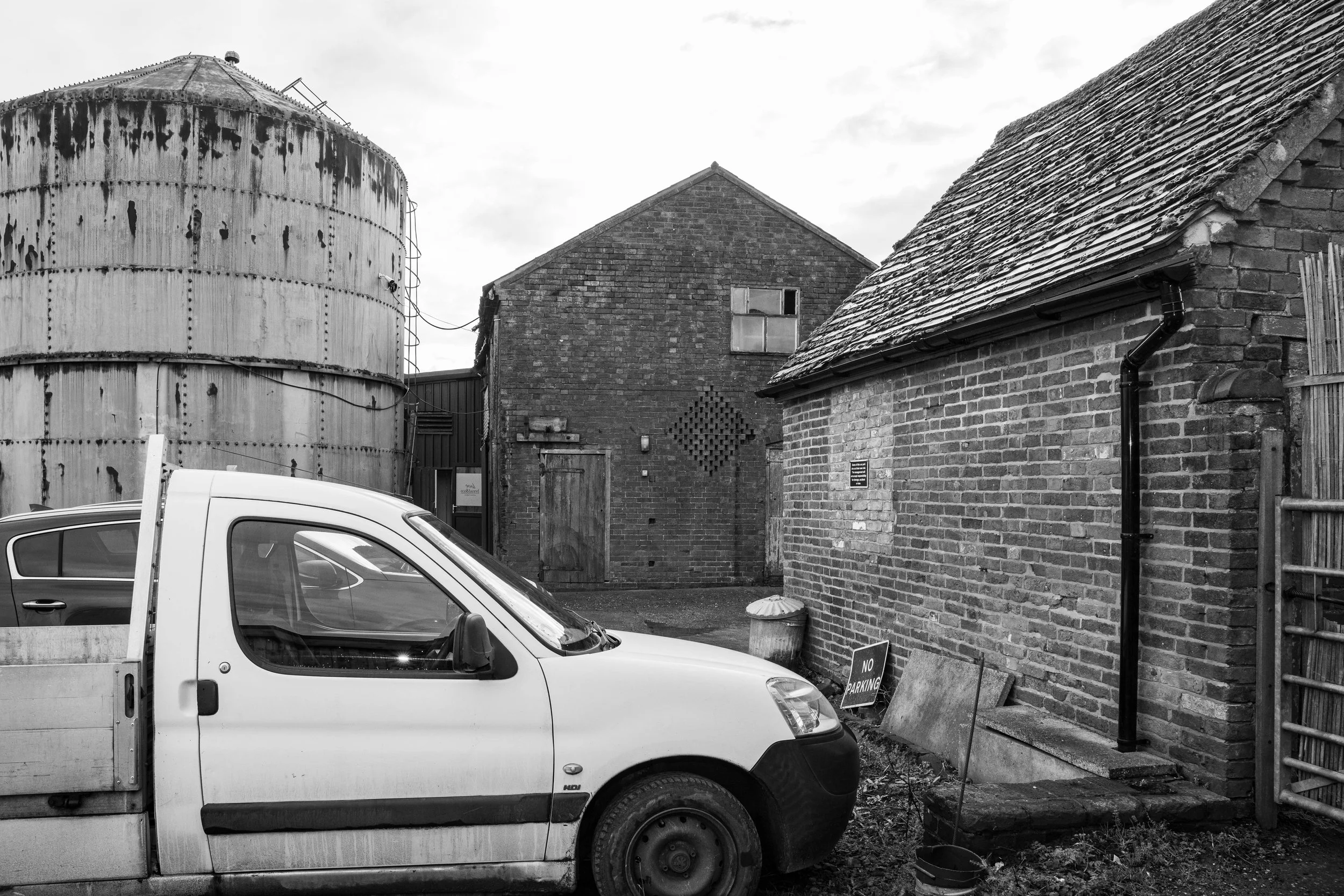Black and white photo of a small brick building with a tiled roof, a large rusty water tank, a white pickup truck, and a "No Parking" sign on the ground.