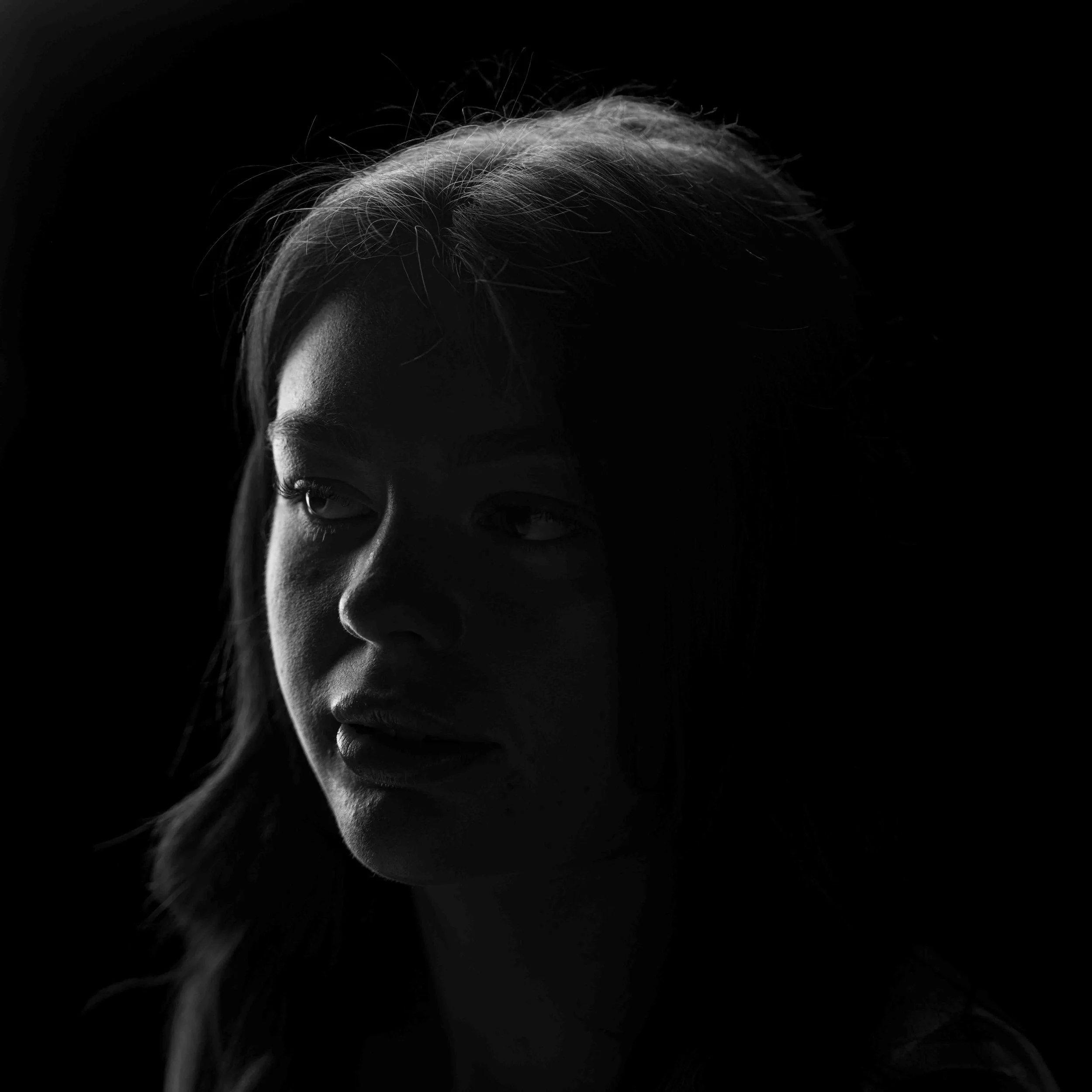 Black and white photo of a woman with light-colored hair styled back, looking downward against a dark background.