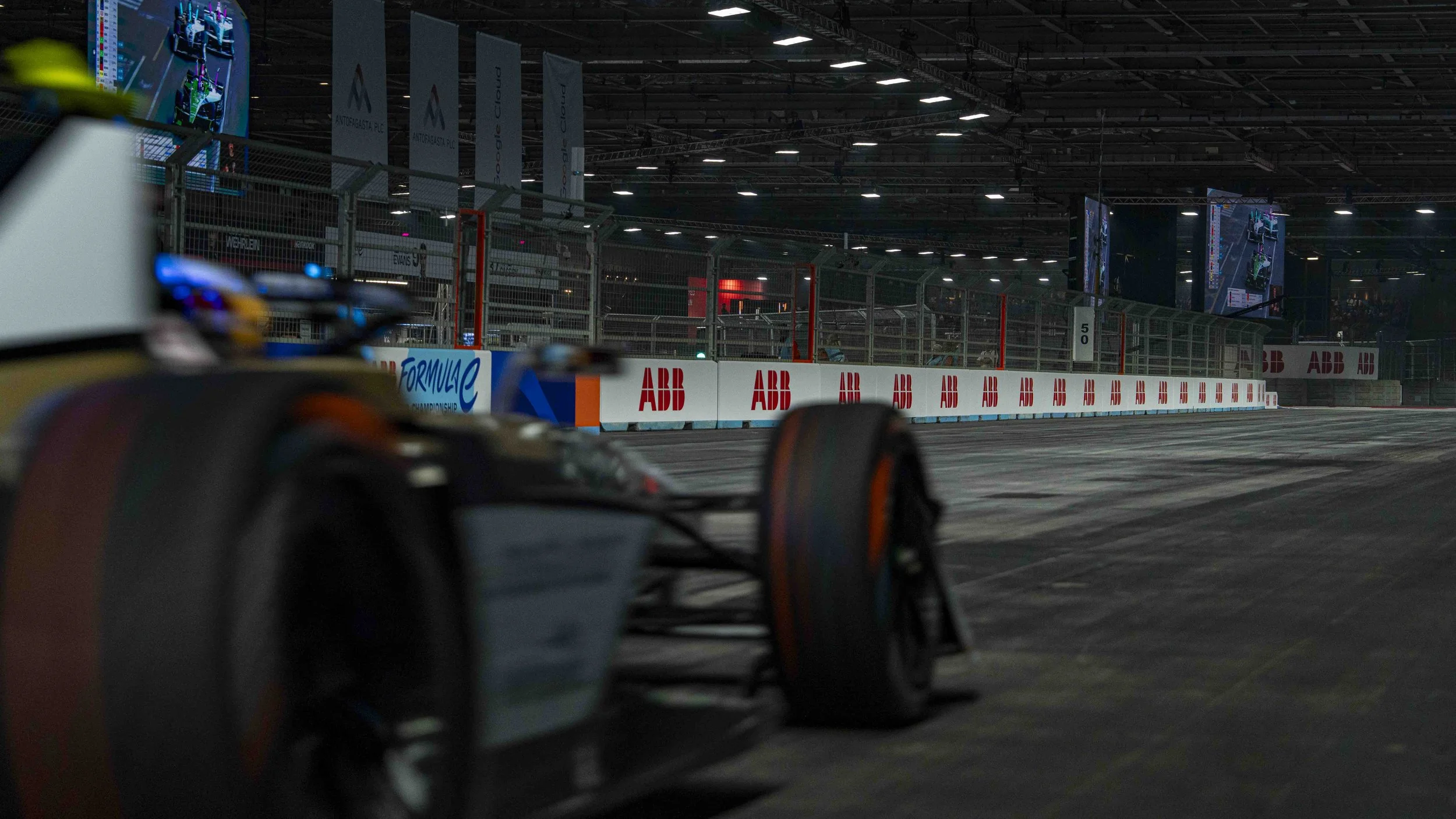 A Formula E race car racing through Excel's indoor track in London with barriers and large screens displaying race footage in the background.