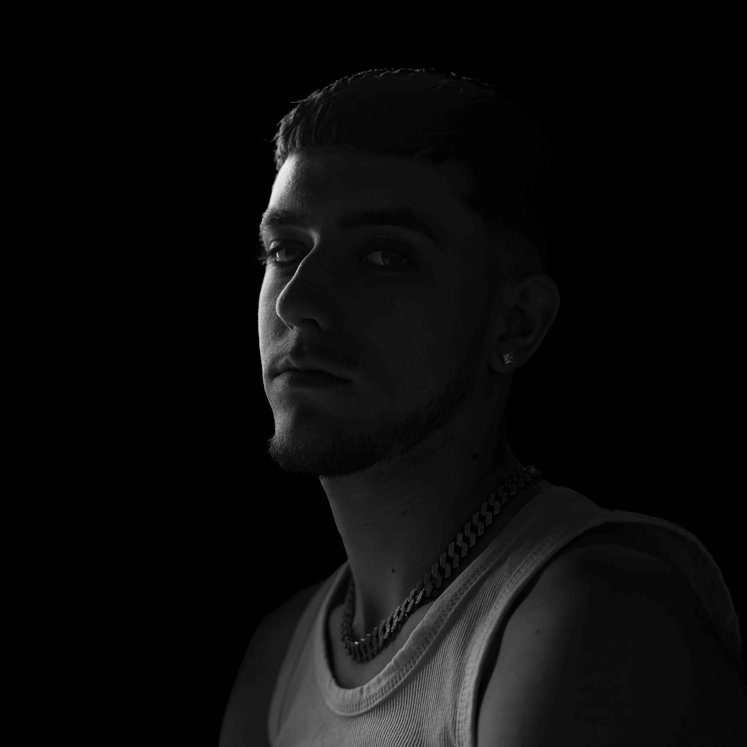 Close-up of a young man's face, with short hair and a serious expression, wearing a chain necklace, collared shirt, and sweater, against a black background.