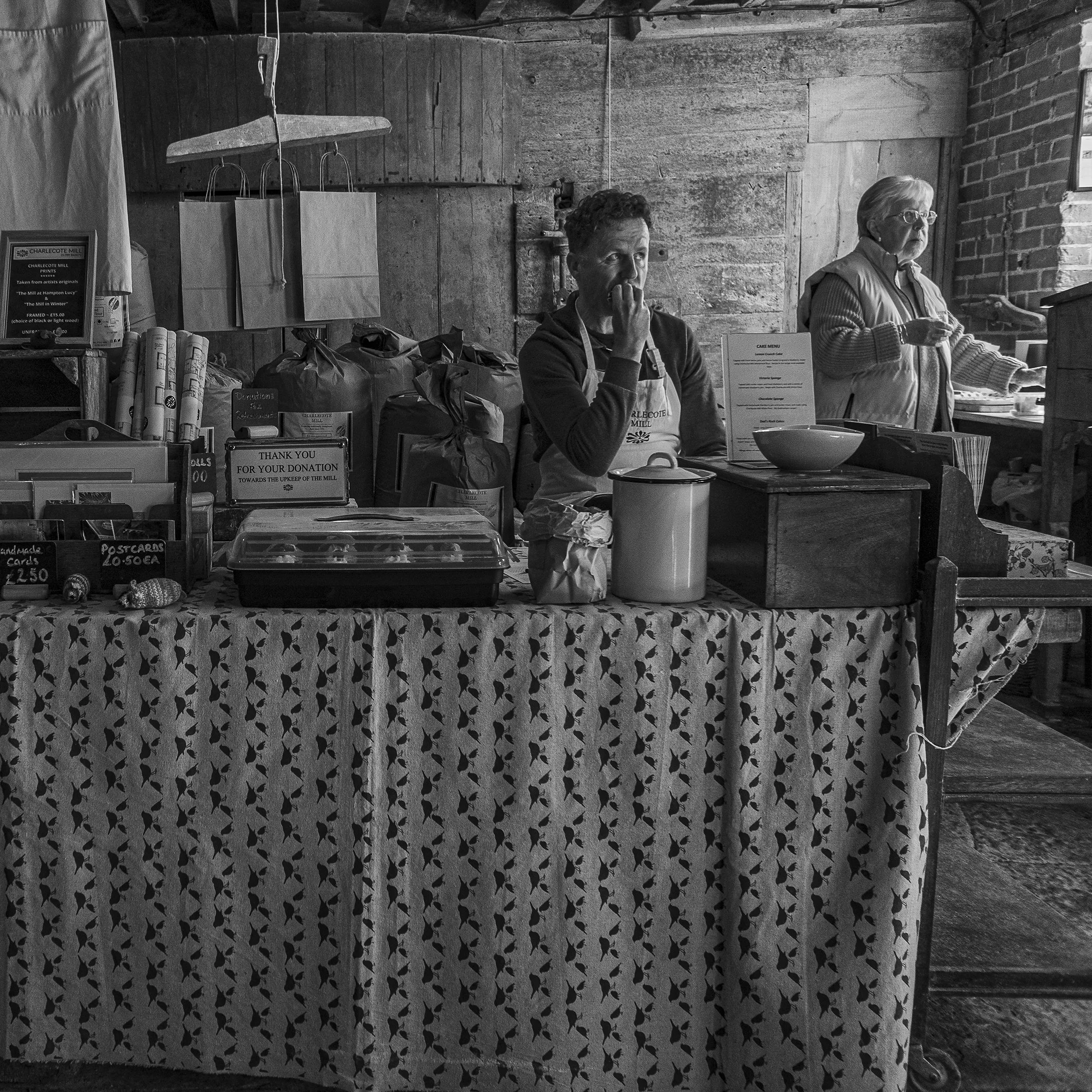A black and white photo of two women sitting behind a table at a market stall. The woman on the left is holding a spoon in her hand and wearing an apron, and the woman on the right is wearing glasses and a striped sweater, with a bowl in front of her