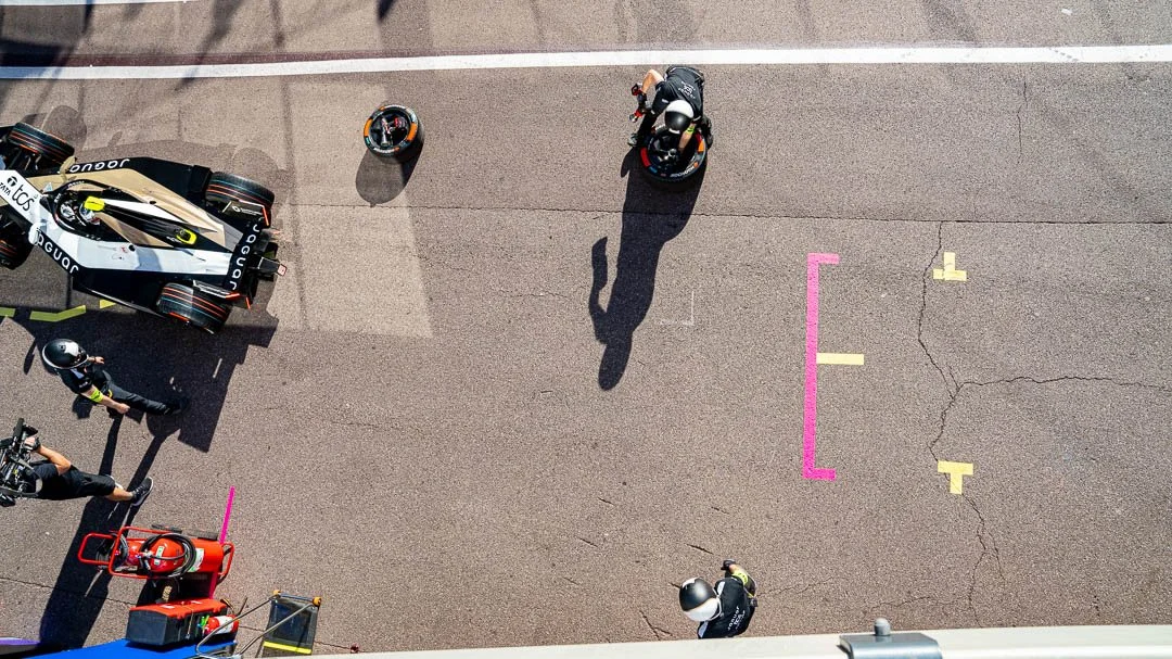 A view from above showing a Formula 1 car in the pit lane, with team members and a motorcycle nearby, marked with pink and yellow lines and markings on the asphalt.