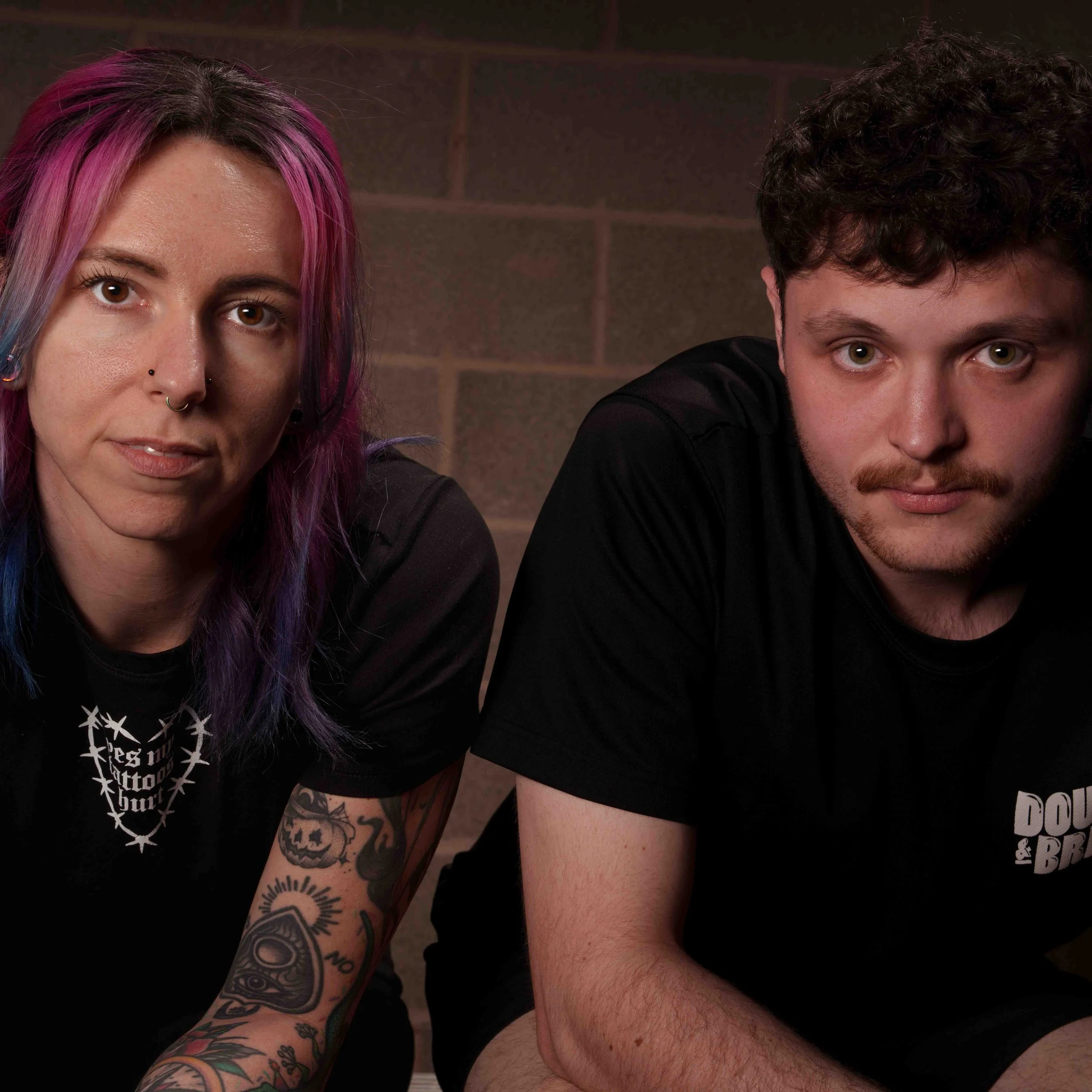 Part of two people with tattoos on arm, with one woman with pink hair and nose piercings, and a man with beard, both leaning forward.