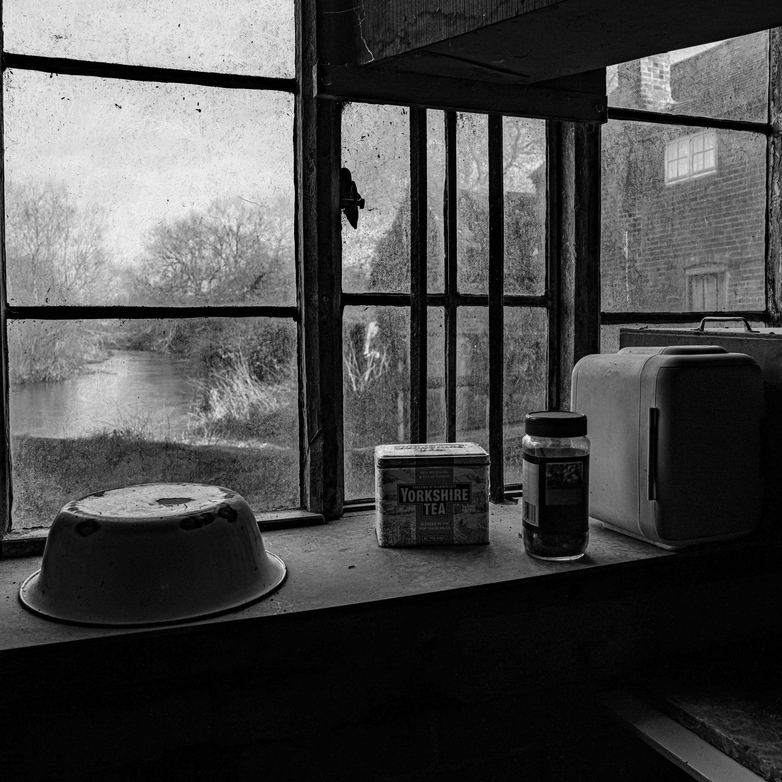Black and white photo of a window sill with a speaker on the left, a box of Yorkshire Tea, a jar, and a toaster on the right, with the view outside showing trees and a brick building.