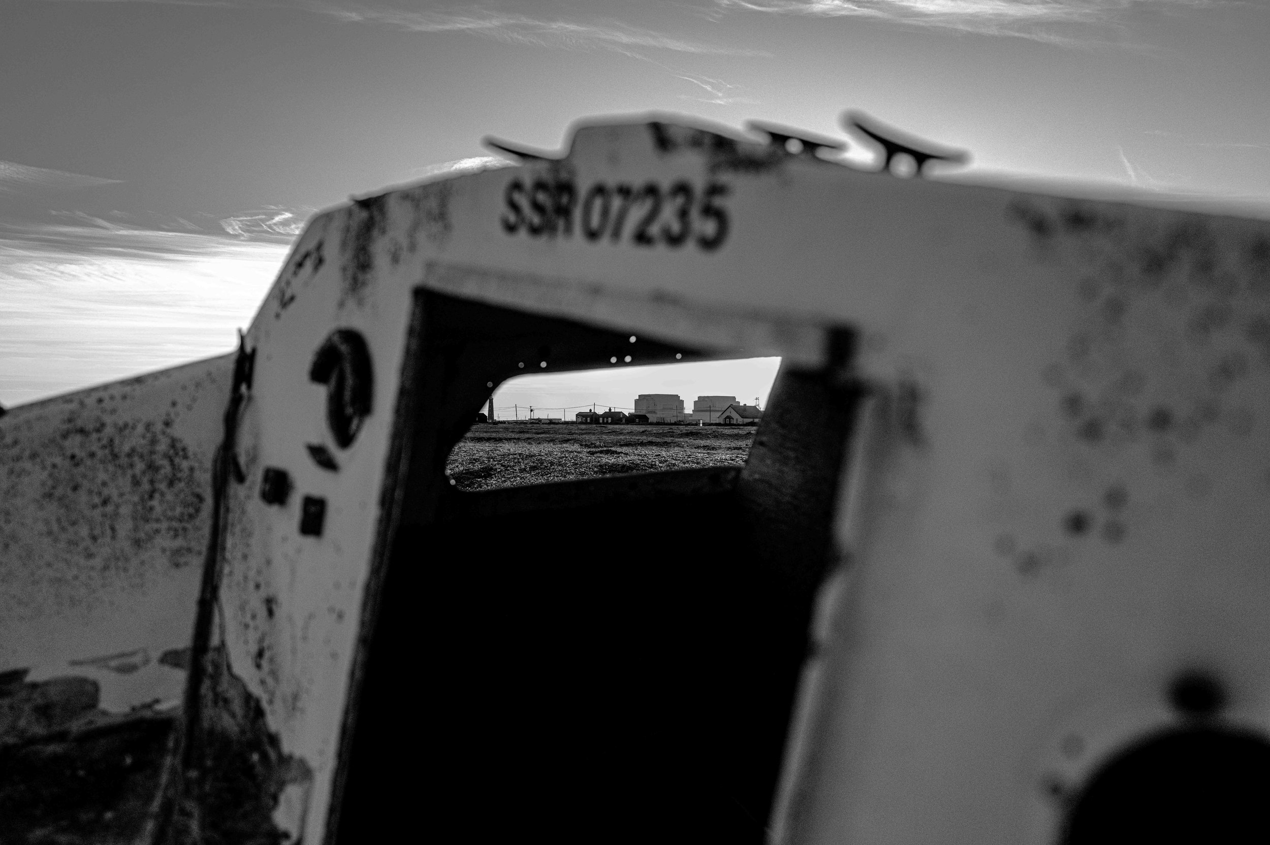 View through a weathered, rusty opening of an abandoned boat, looking toward a distant landscape with a few buildings and power lines under a cloudy sky in Dungeness