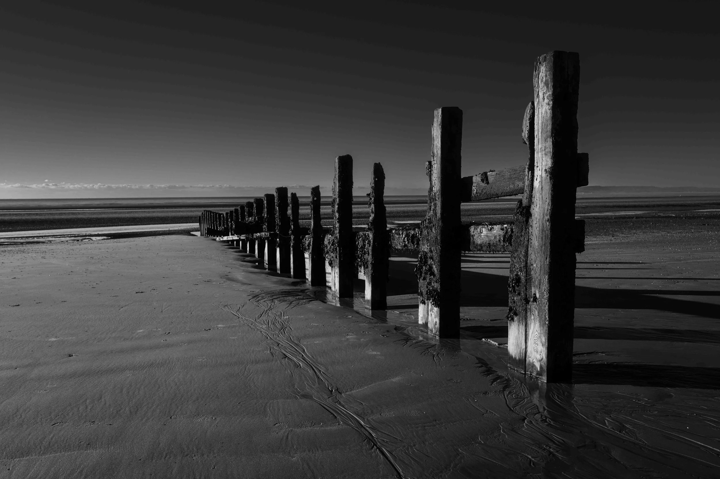 Black and white photo of a weathered groynes barrier along a sandy beach, with the shoreline and ocean in the background under a clear sky.