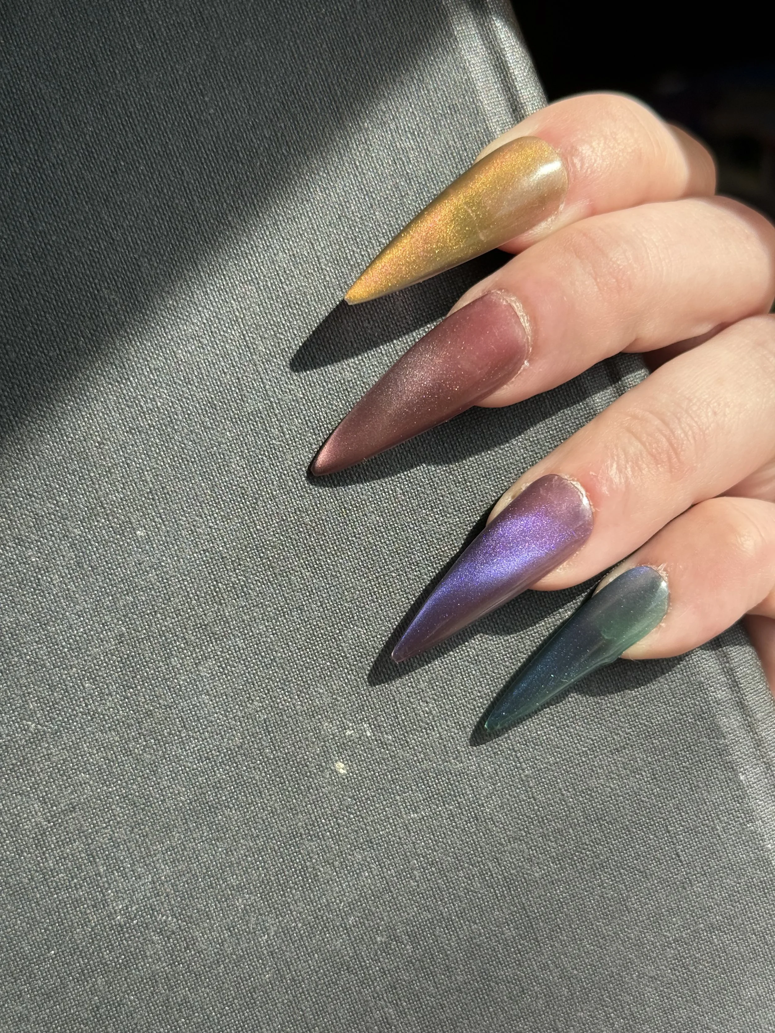 Close-up of a hand with colorful, metallic acrylic nails in yellow, pink, purple, and green, resting on a textured gray surface.