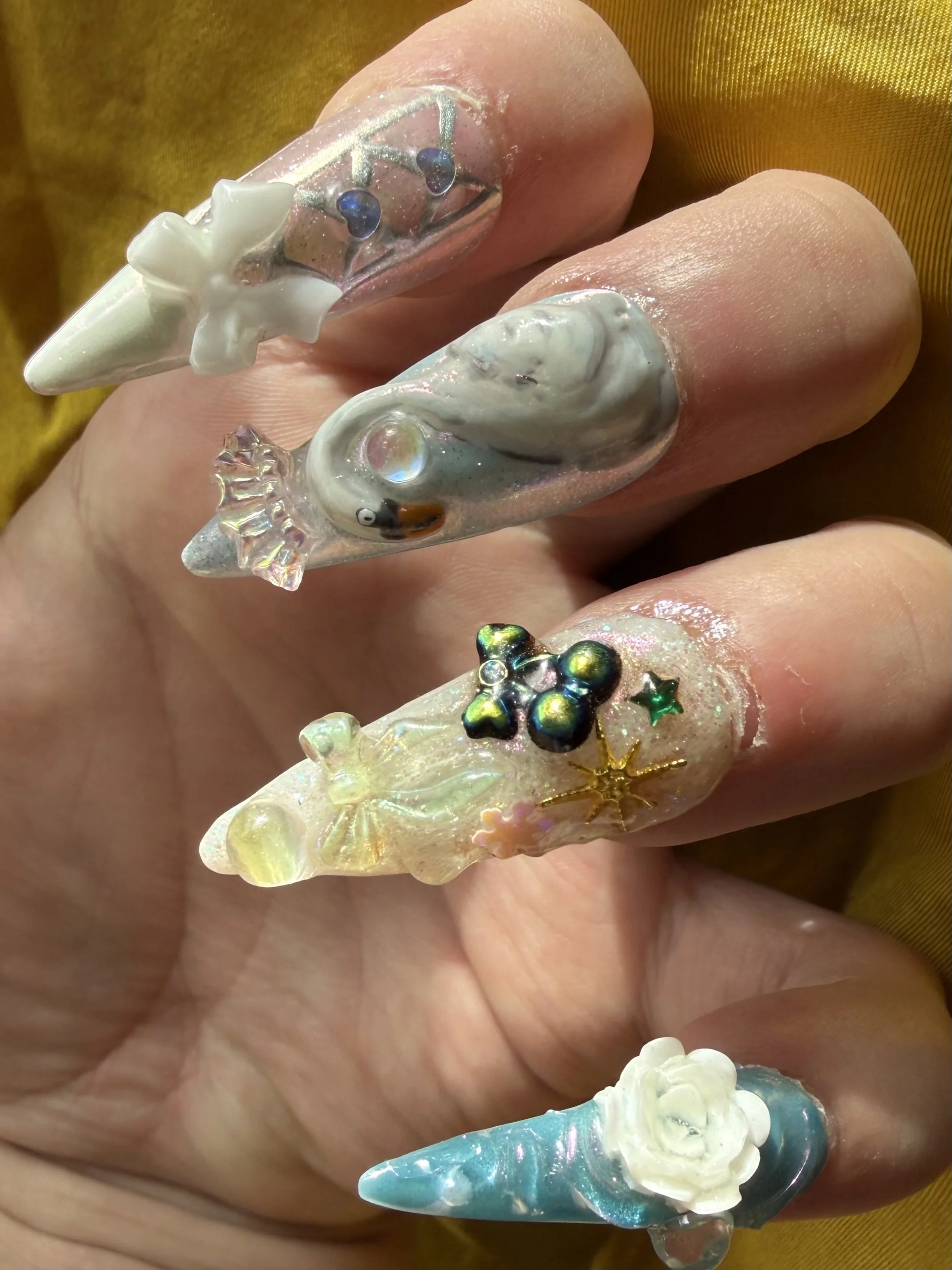 Close-up of a hand with elaborate 3D nail art featuring unicorn horn designs, pearls, and holographic embellishments.