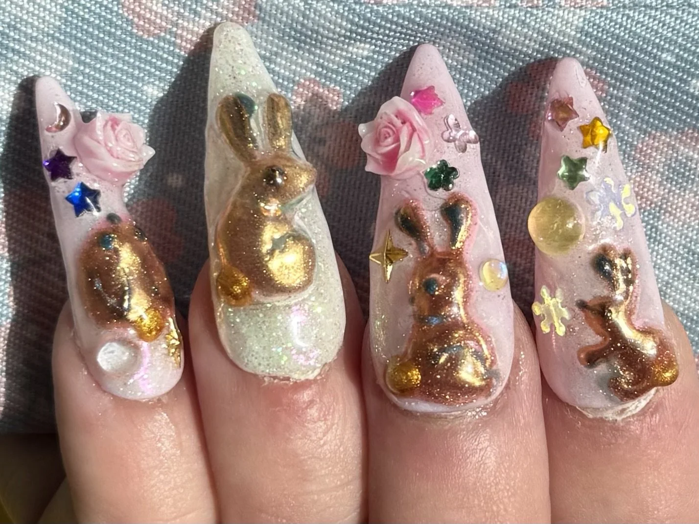Close-up of a hand with decorated nails featuring 3D bunny figures, pink roses, and colorful star-shaped glitter on a transparent base, with a silver textured background.
