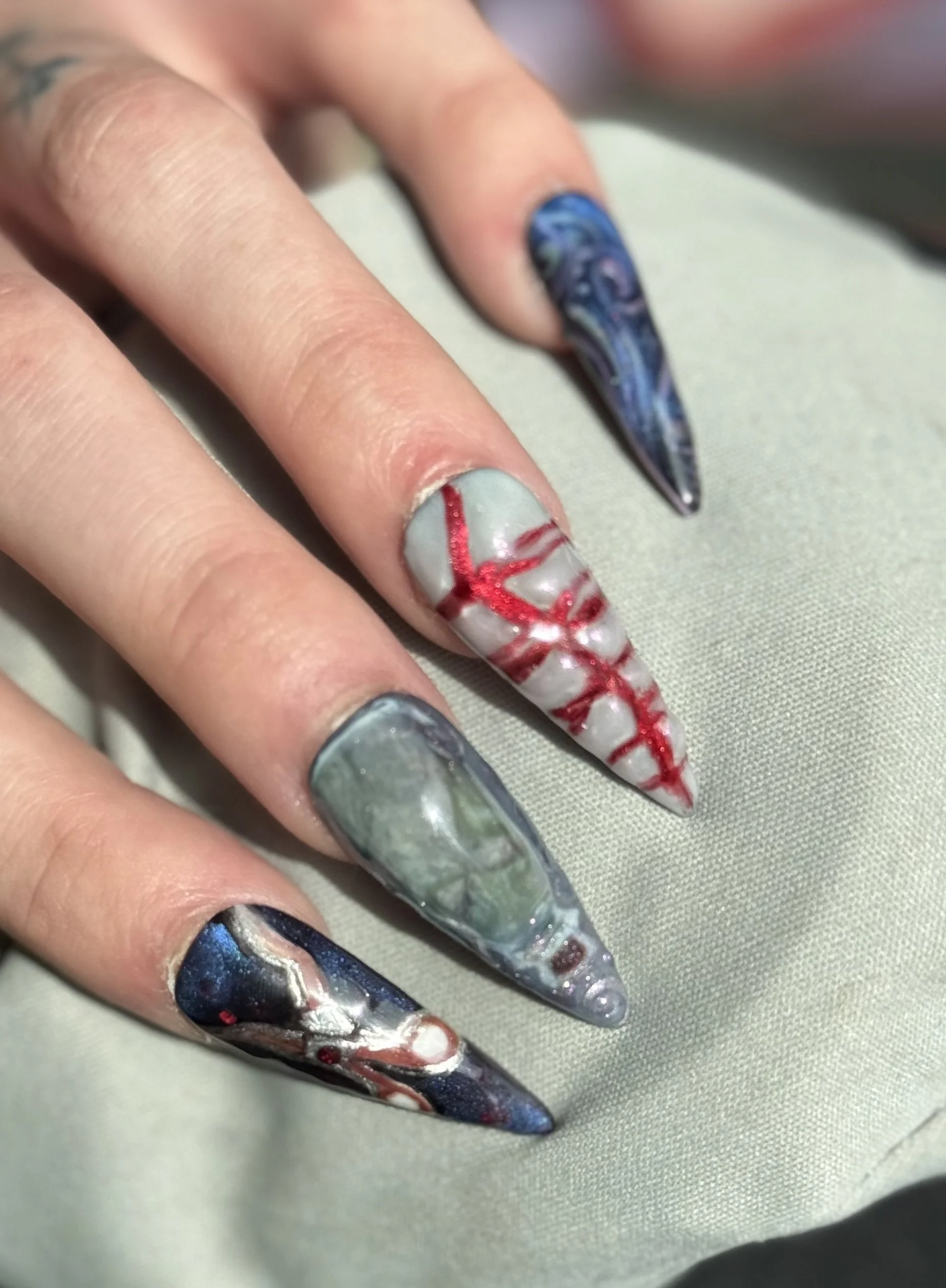 Close-up of a hand with long, pointed, multicolored gothic-style nails featuring artistic designs like galaxy, marble, and horror-inspired effects.