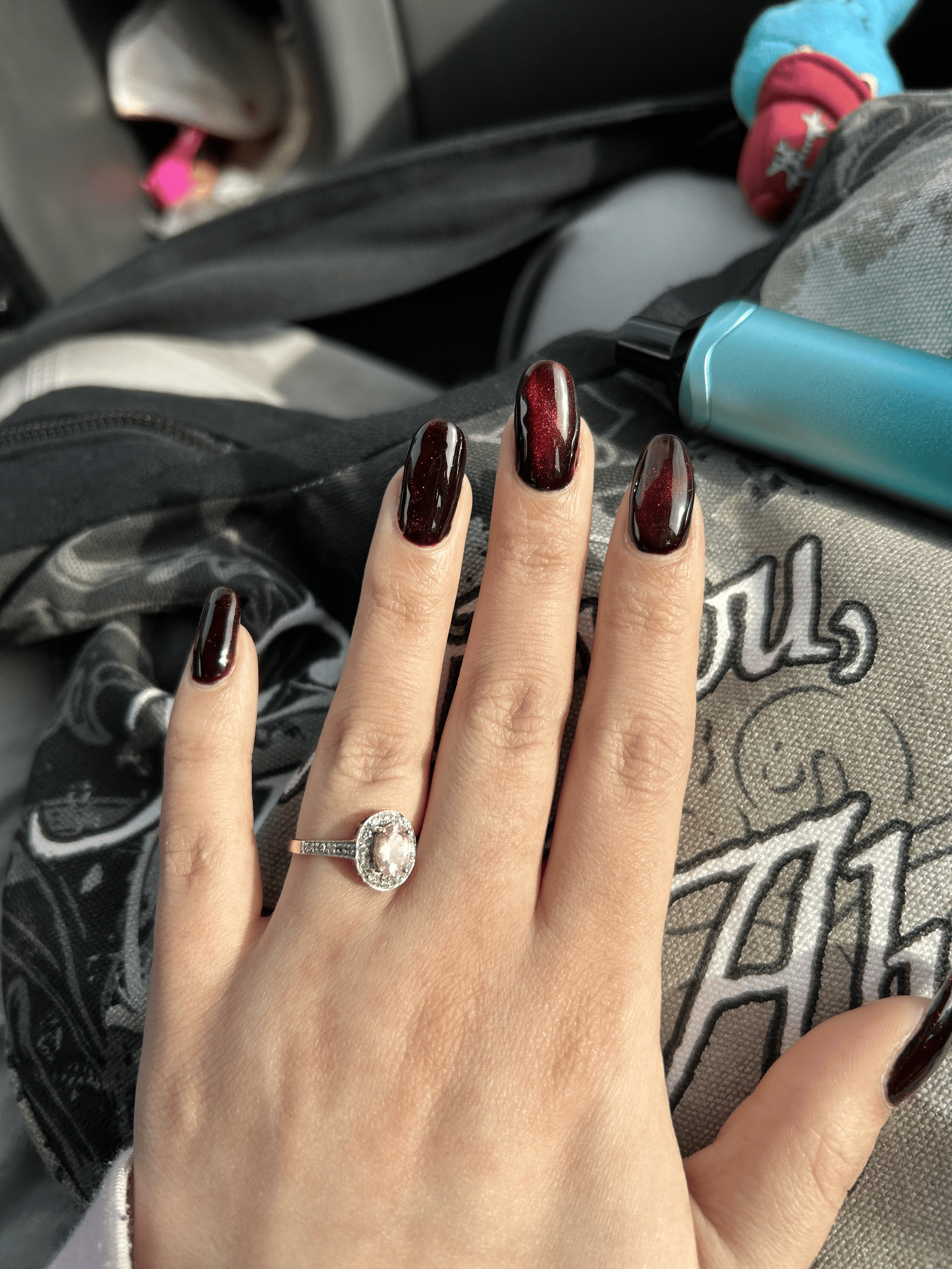 A hand with dark red, glossy nails and a large oval diamond ring on the ring finger resting on a black and gray backpack with text and a graphic.