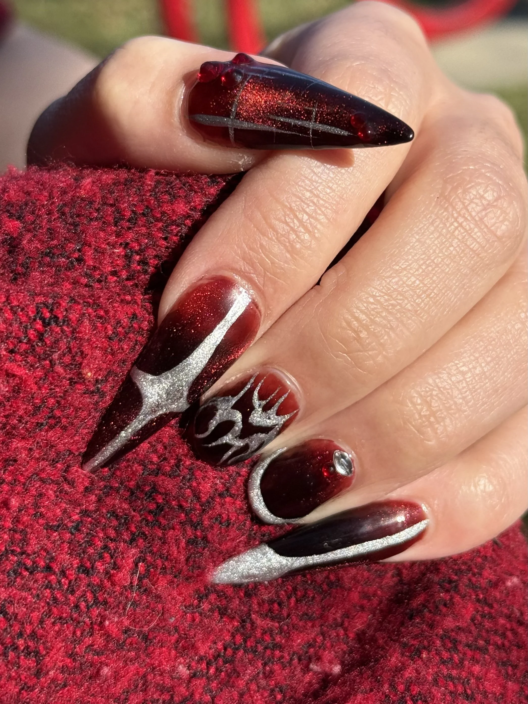 Close-up of a hand with long, pointed nails painted in black and red with silver accents and rhinestones, resting on a red textured fabric.