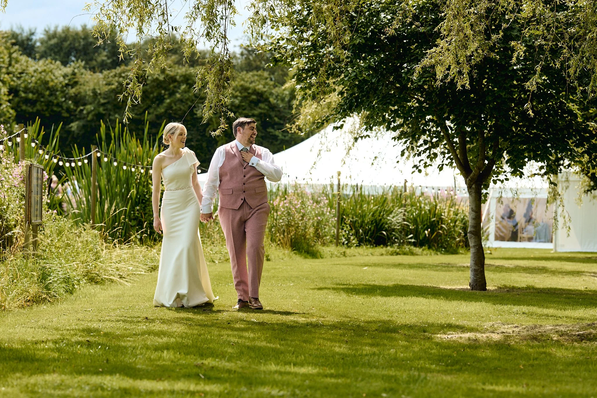 A bride and groom holding hands and walking outdoors on a grassy area during their wedding, smiling and enjoying the sunny day.