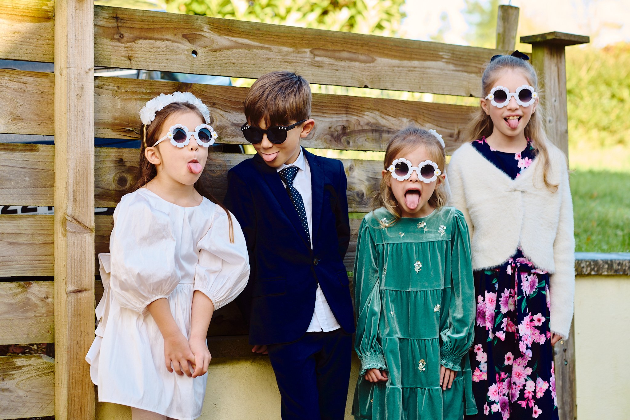 A group of five children standing outdoors in front of a wooden fence, wearing colorful dresses and sunglasses, making playful faces, and sticking out their tongues.