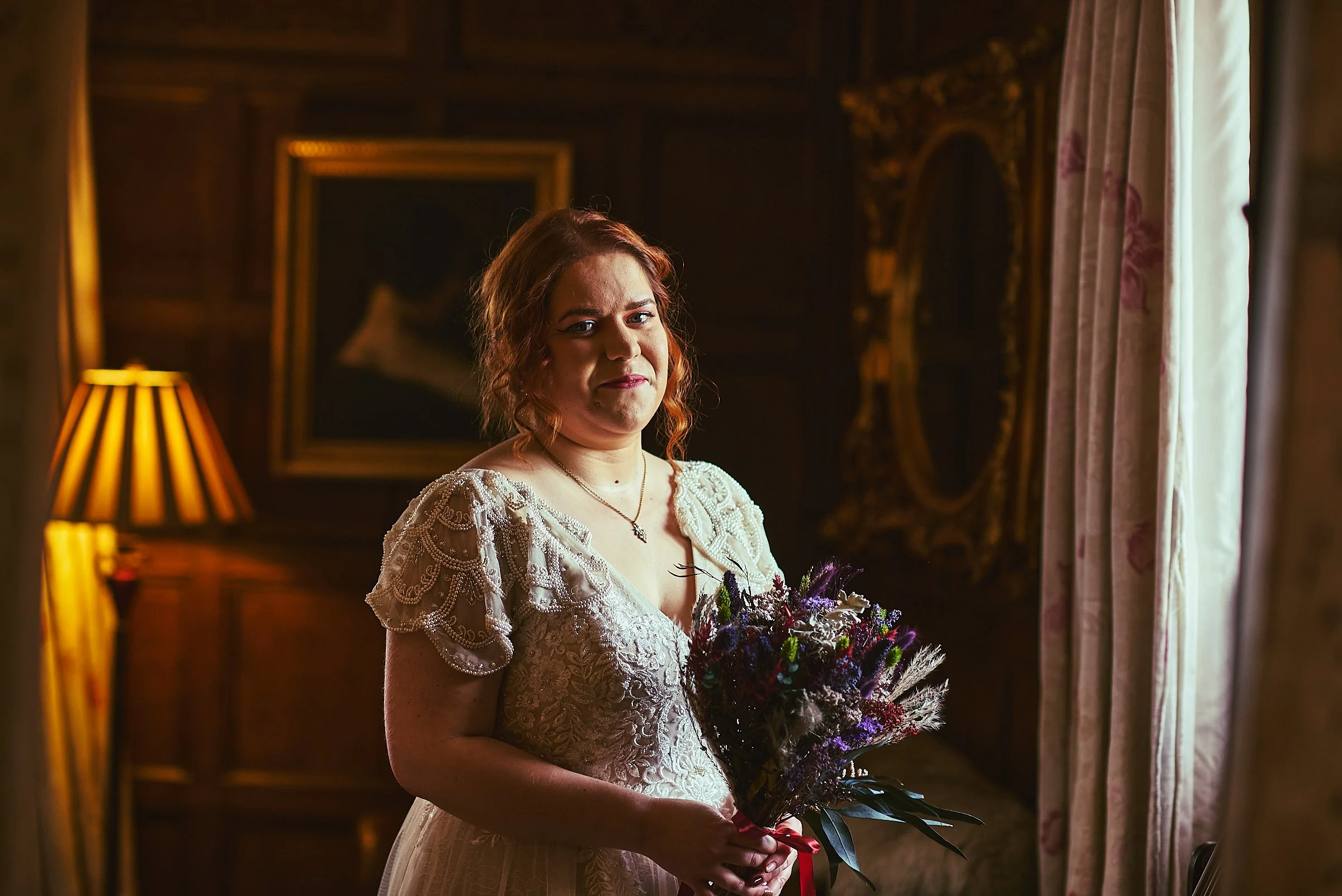 A woman with red hair dressed in a lace wedding gown holding a bouquet of flowers in a dimly lit, elegant room.