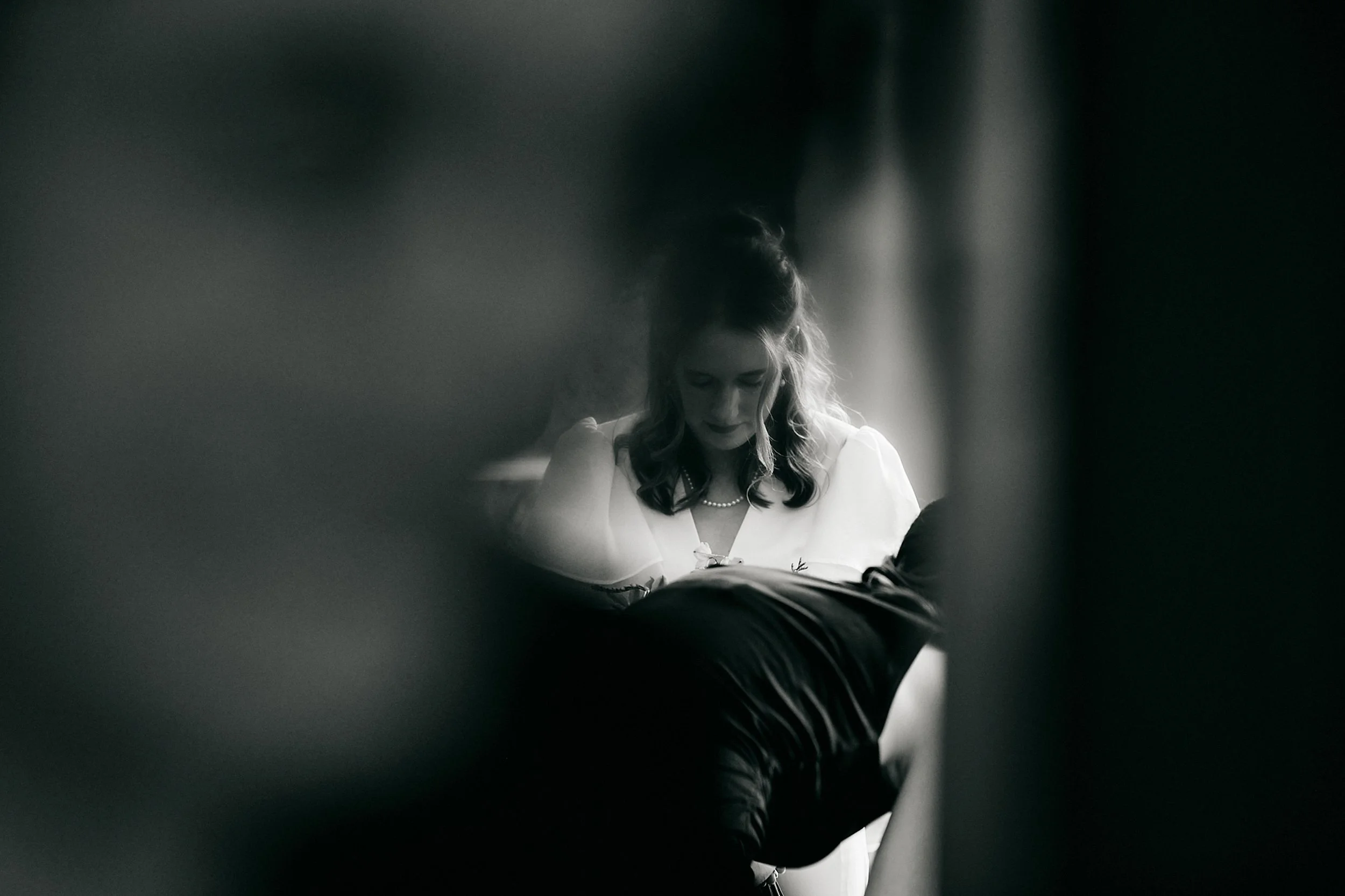 A woman with long hair wearing a pearl necklace reading a book, viewed through a narrow opening or gap.