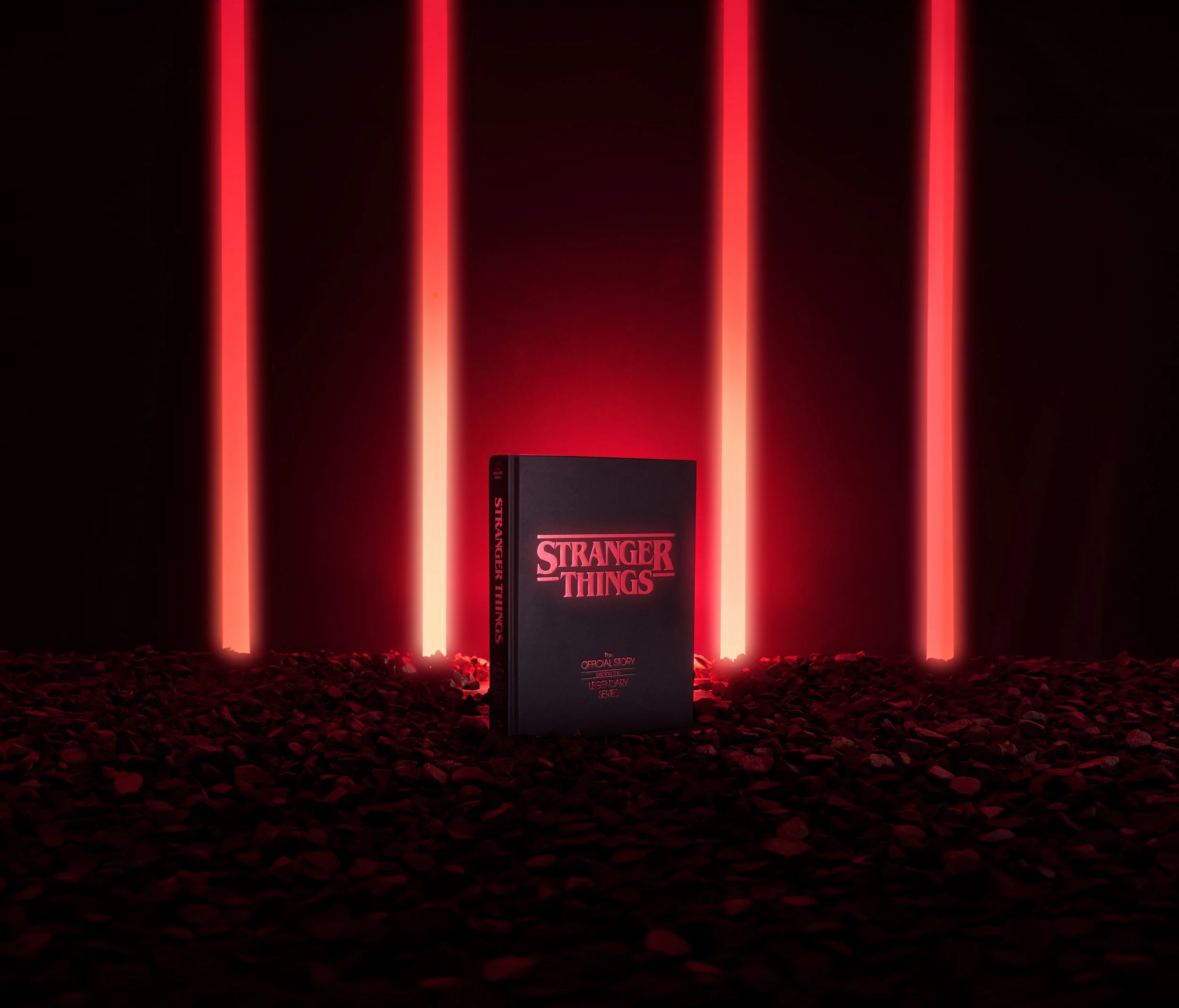 A black book titled 'Stranger Things' stands upright on a ground covered with small rocks, illuminated by four vertical red neon lights against a dark background.