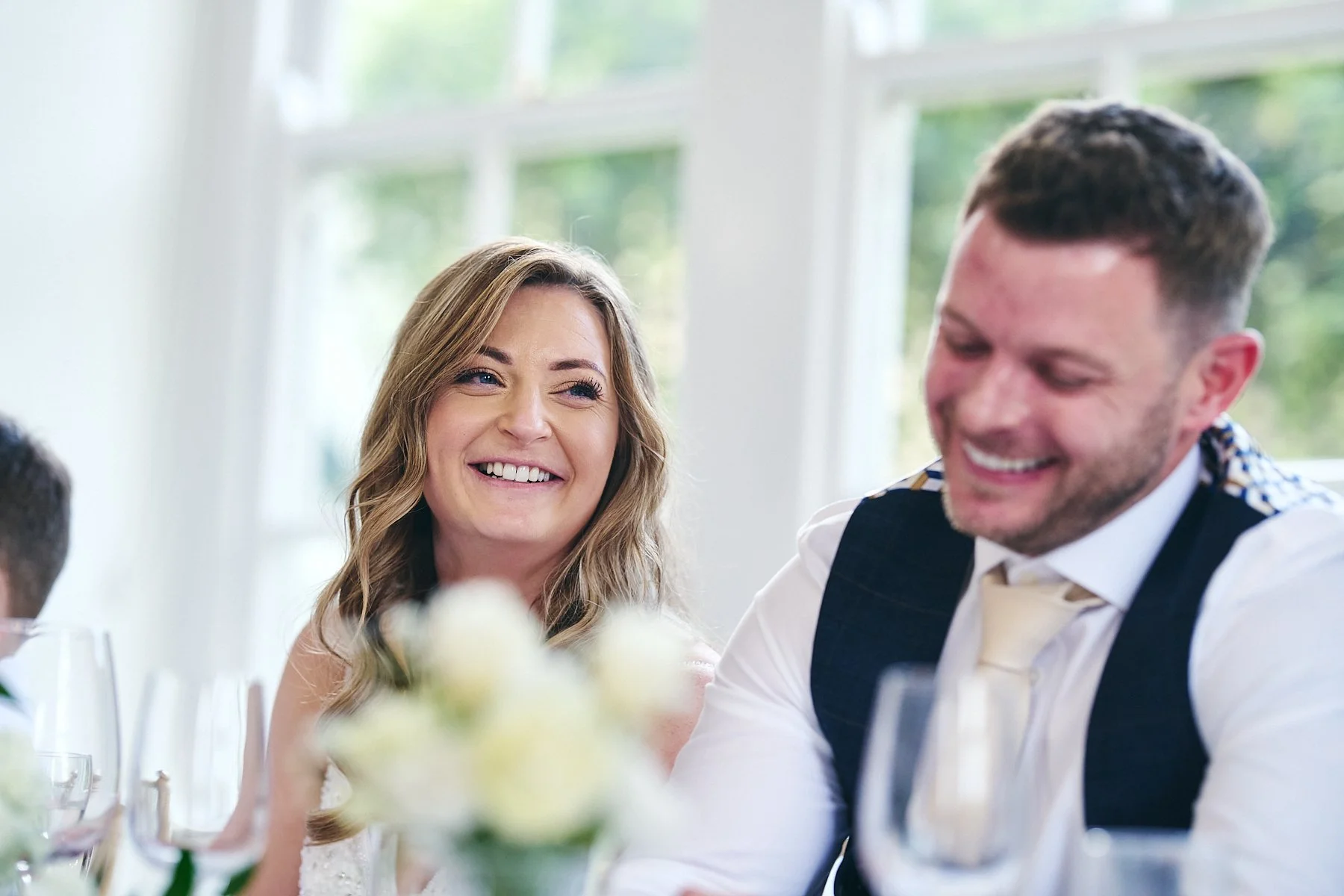 A woman and a man smiling and laughing at a wedding reception table with glasses and flowers.