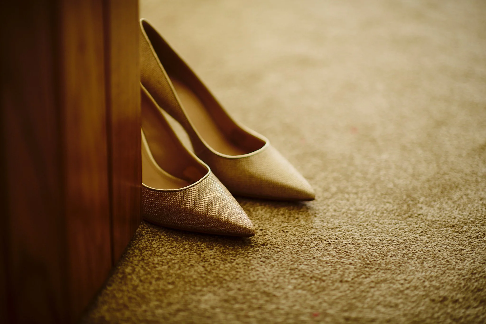 A pair of beige high-heeled shoes placed on a carpeted floor, with a wooden piece of furniture on the left side of the image.