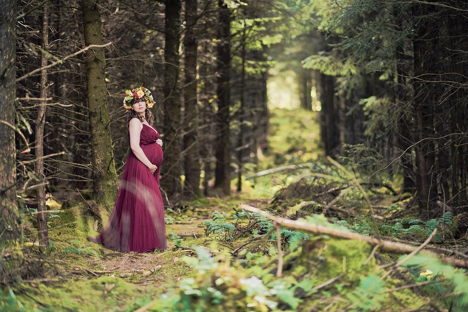 A pregnant woman in a long, flowing maroon dress standing in a dense forest with trees and green ferns, wearing a large floral crown.
