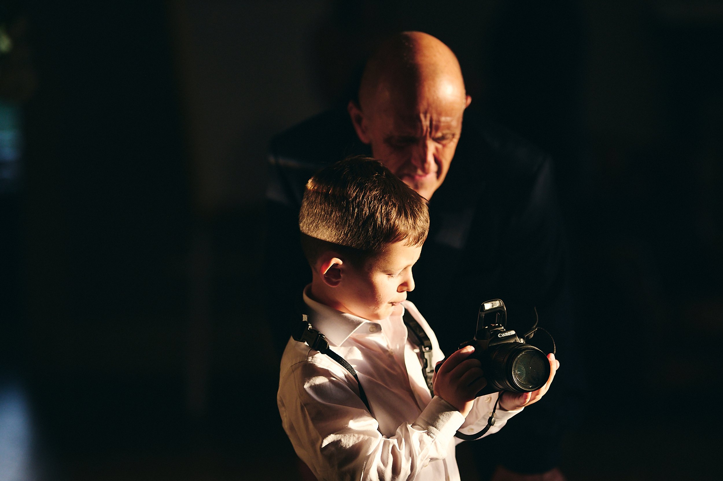 A young boy with short brown hair and a white shirt holding a Canon camera, looking at the camera, with an older man with a bald head and dark clothing leaning over him in a dark setting.