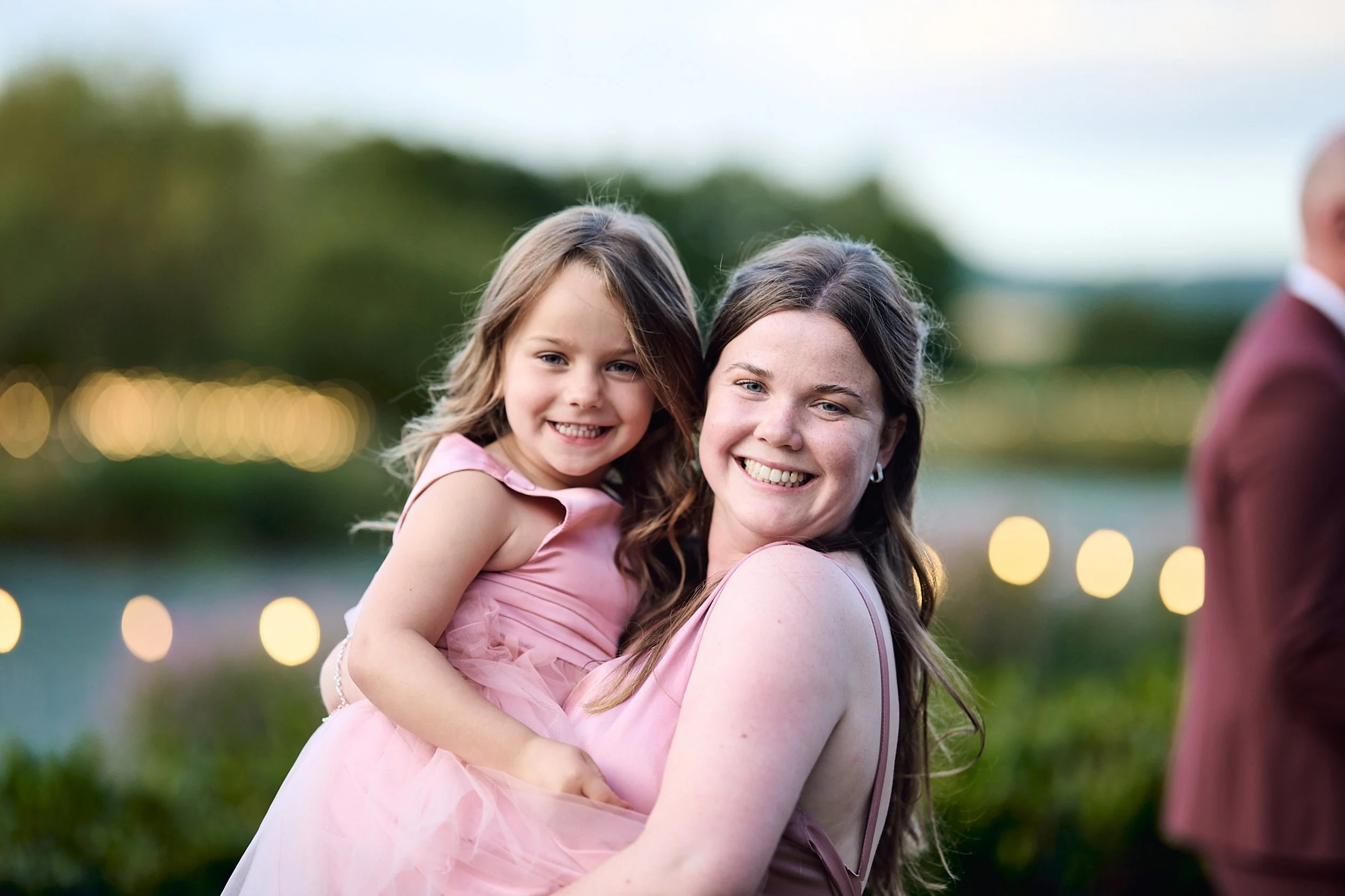 A woman and a girl smiling, with the girl sitting on the woman's shoulders outdoors during evening or dusk, with blurred background lights and greenery.