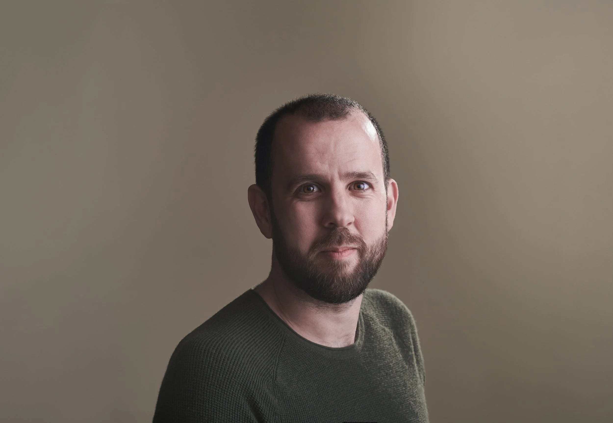 A man with short dark hair and a beard, wearing a dark sweater, smiles softly at the camera against a plain, beige background.