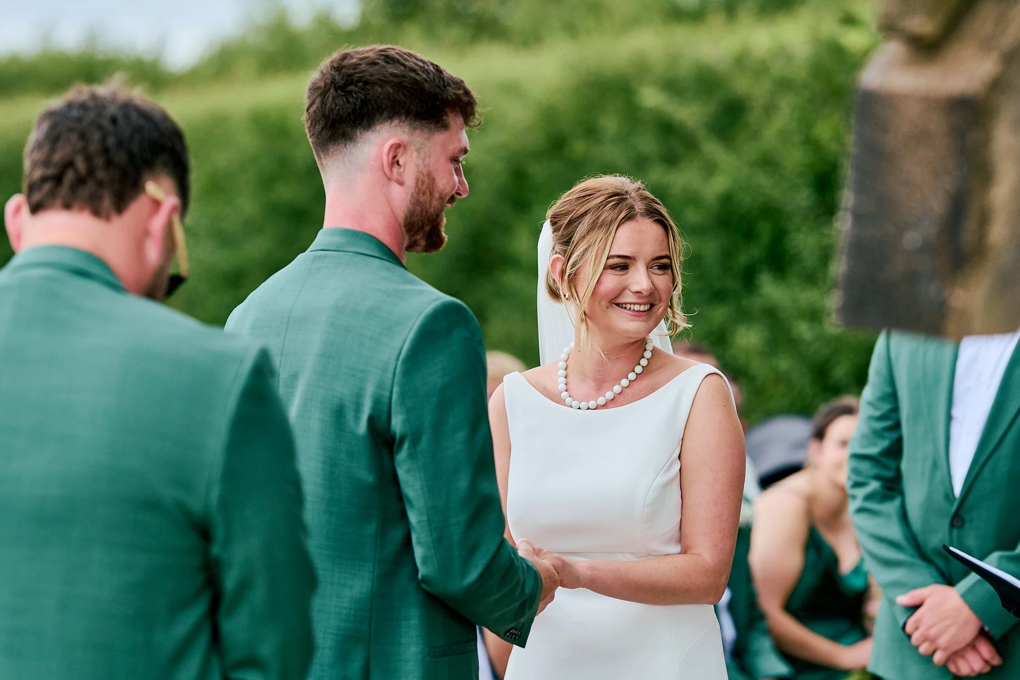 A bride and groom exchanging vows during an outdoor wedding ceremony, with the bride wearing a white dress and necklace, and the groom in a green suit, while an officiant stands nearby.