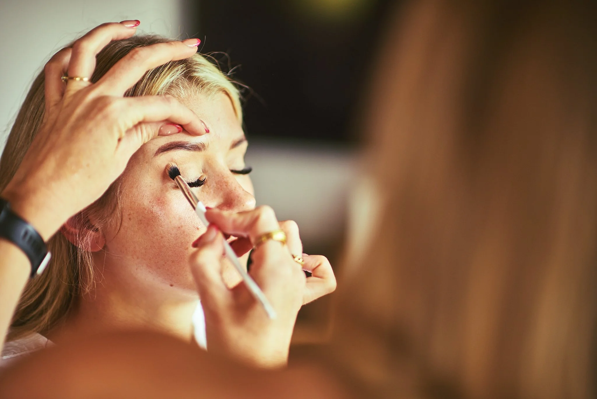 A person with light brown hair having makeup applied near their eye, with another person using a brush to put eyeshadow or eyeliner on them.