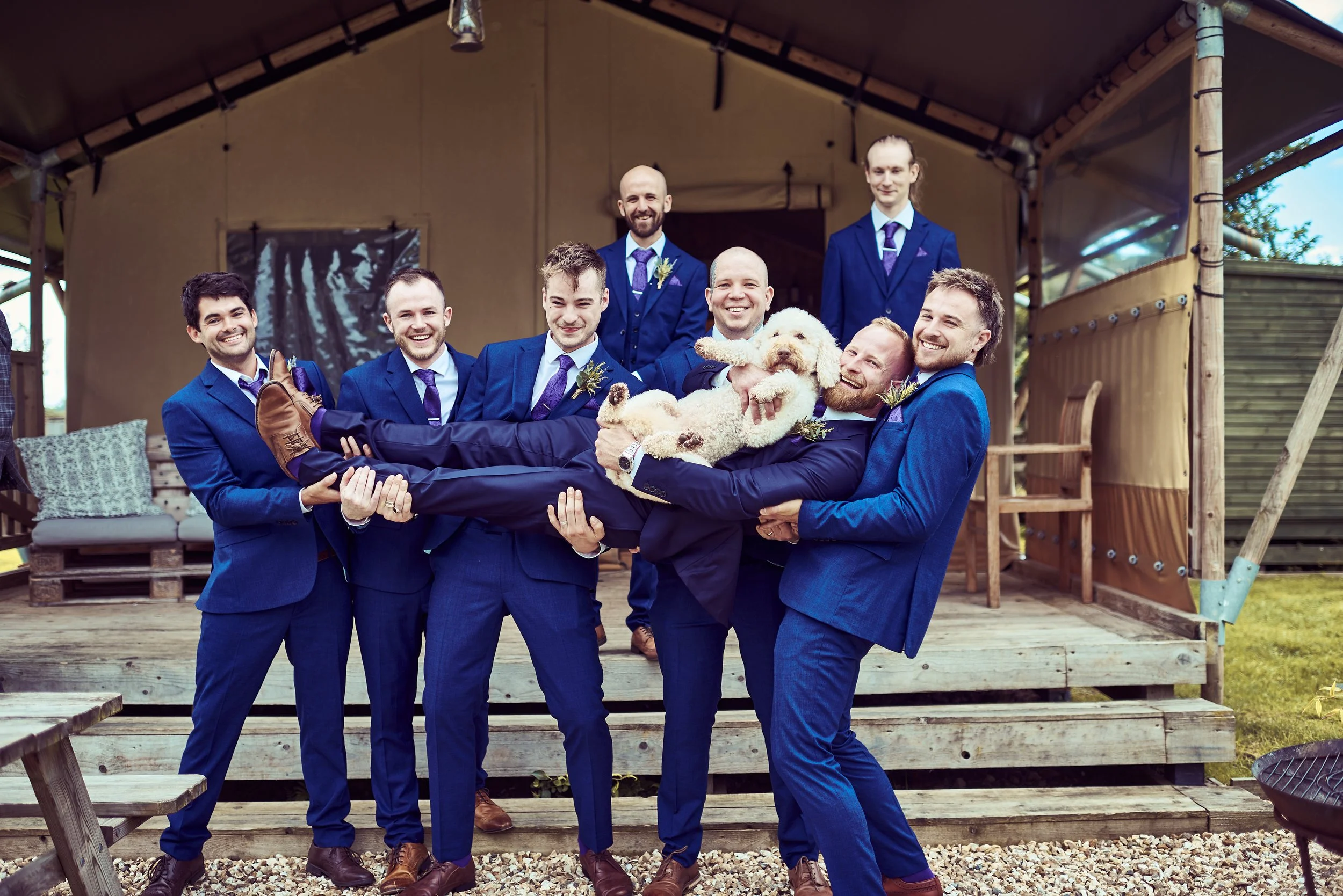 A group of nine men in matching blue suits and purple ties, some with boutonnières, are smiling and posing for a photo while holding a small dog. They are standing on a wooden deck outside a building with a tent-like structure and wooden pallets, wit