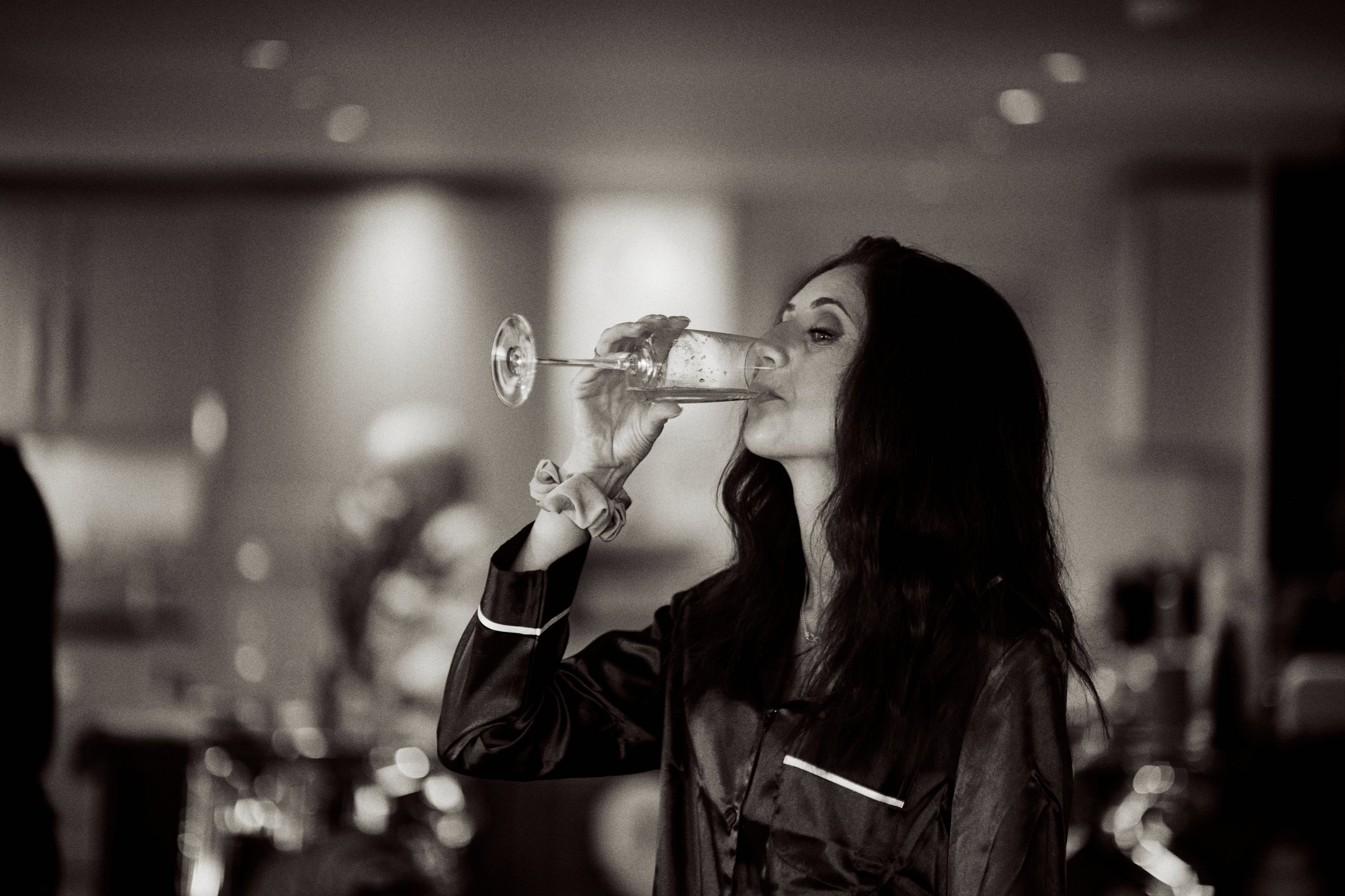 A woman with long dark hair drinking from a wine glass in a dimly lit indoor setting.