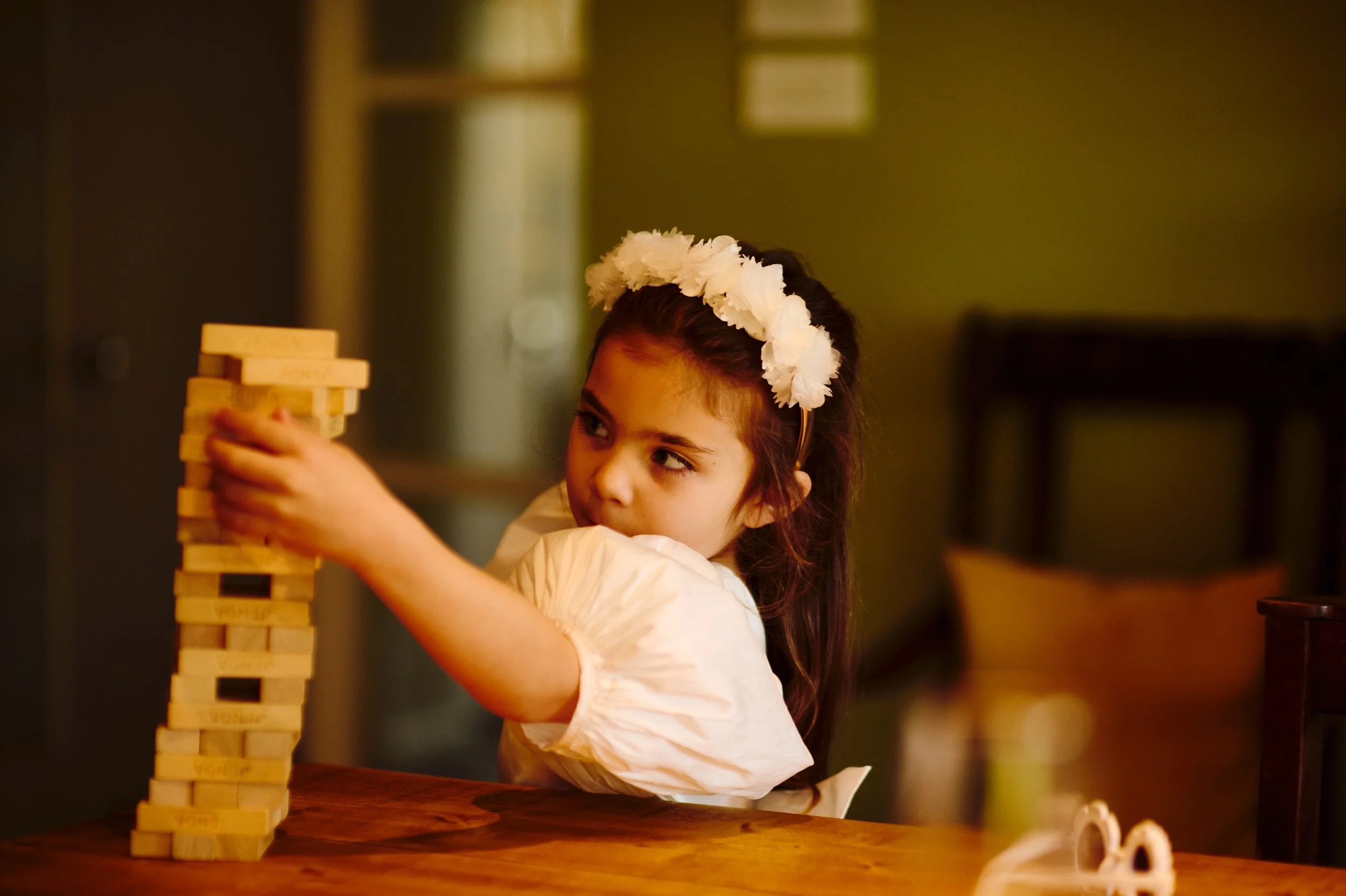 Young girl with a white flower headband stacking wooden blocks on a table.