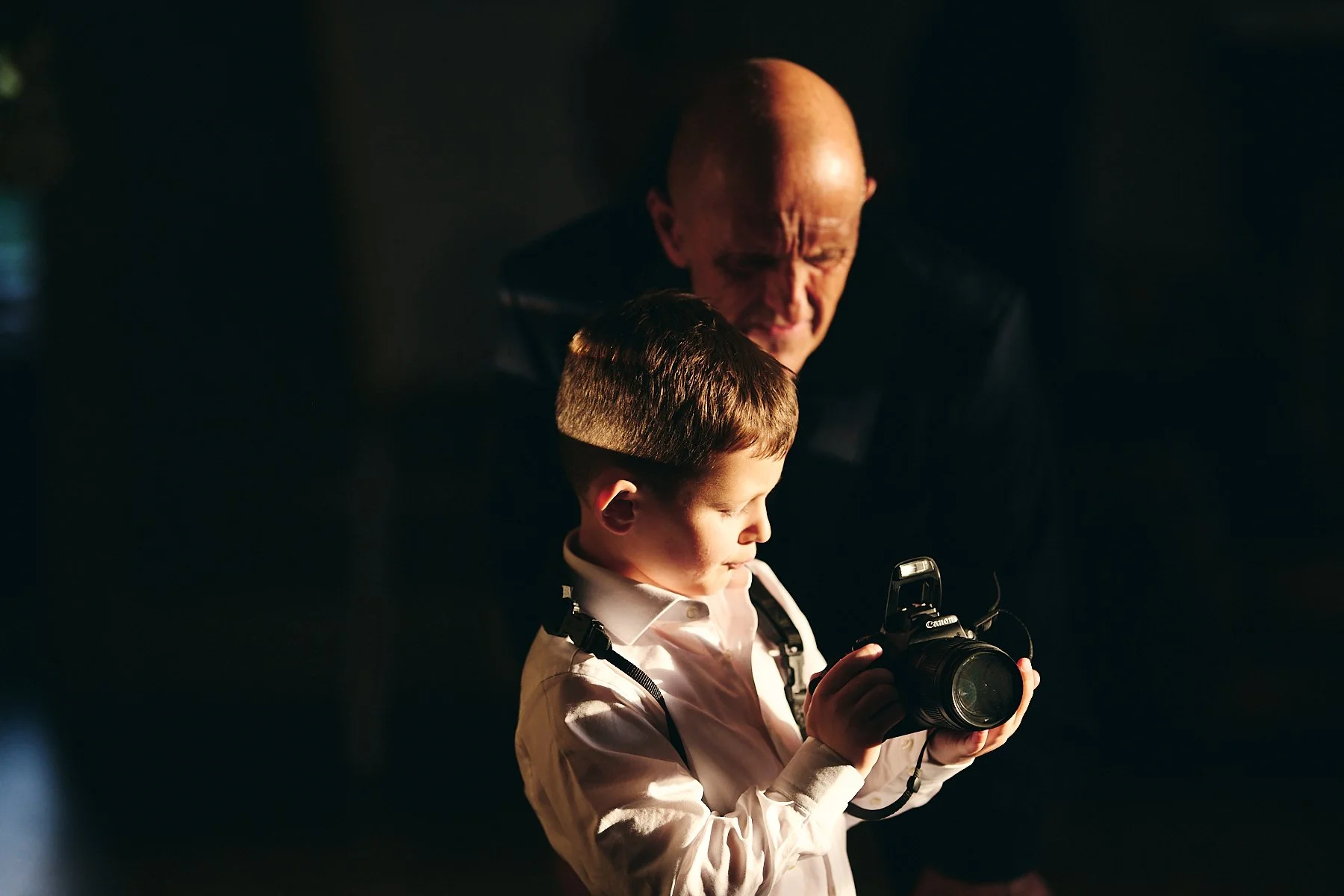 A young boy in a white shirt looking at a camera, with an older man standing behind him, both focused on the camera, in a dark setting.