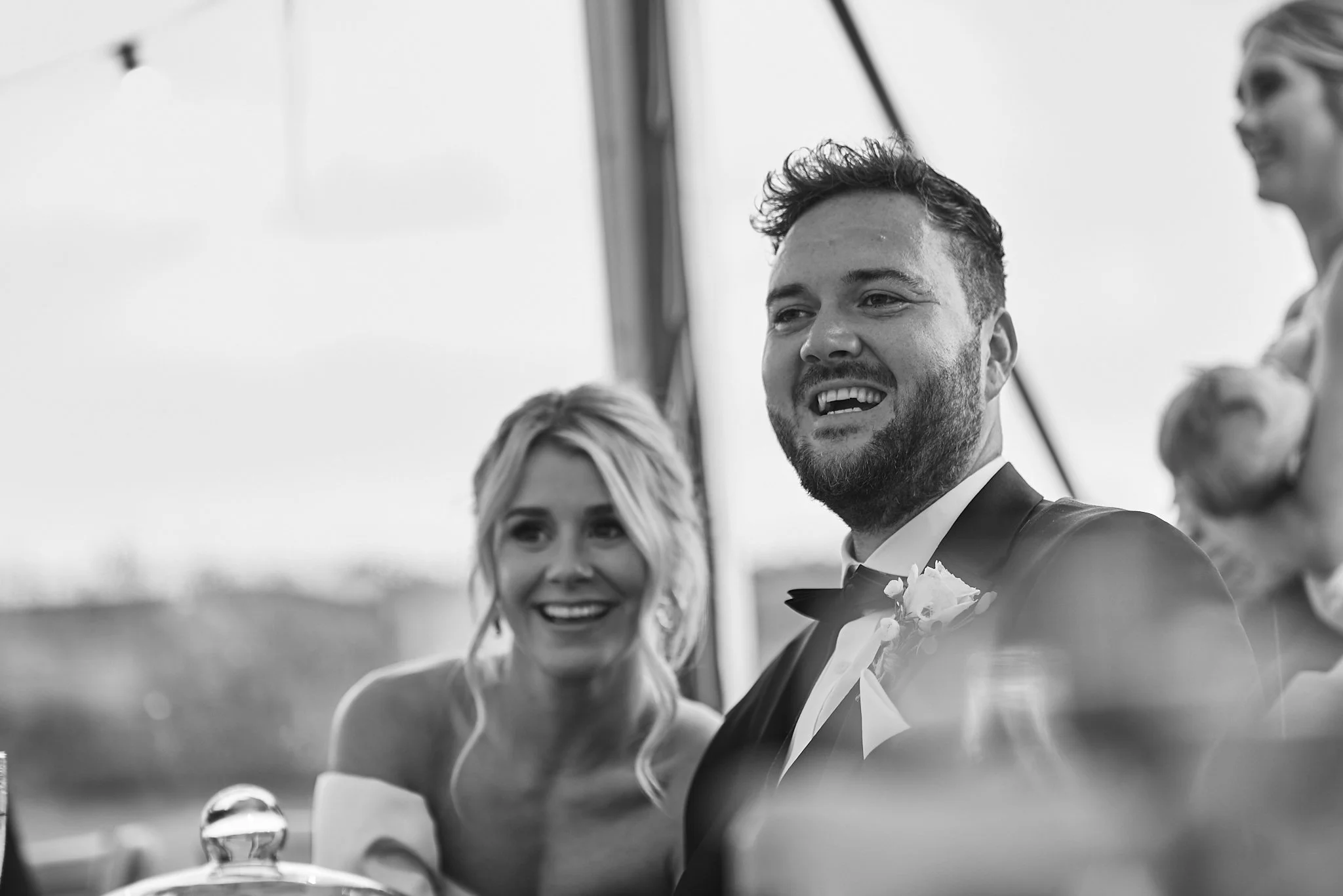 Black and white photo of a smiling man in a tuxedo with a boutonniere, and a woman with long hair and earrings, at an outdoor wedding or formal event.