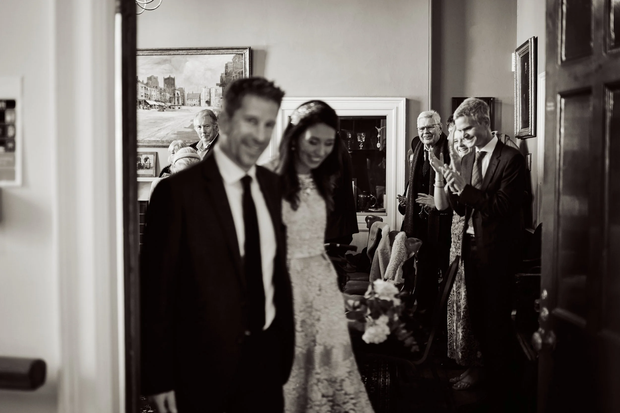 People gathered in a room, smiling and talking; the photo is framed through a doorway, with a painting and shelves visible on the walls.