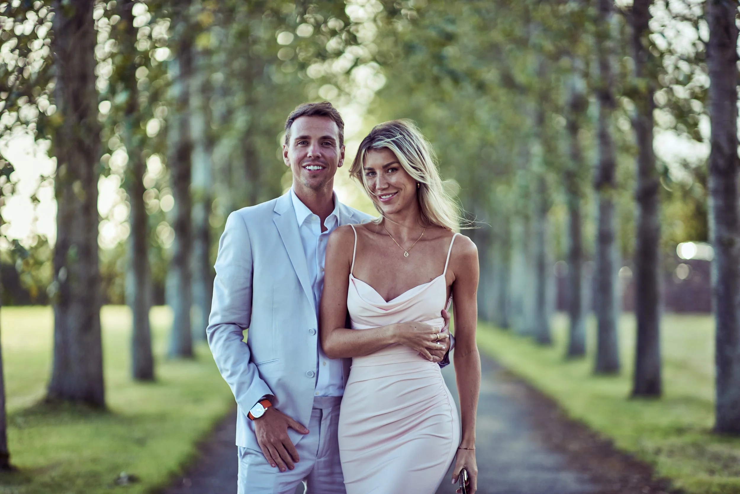 A smiling couple, a man in a light suit and a woman in a light dress, standing close together outdoors in a park with trees in the background.