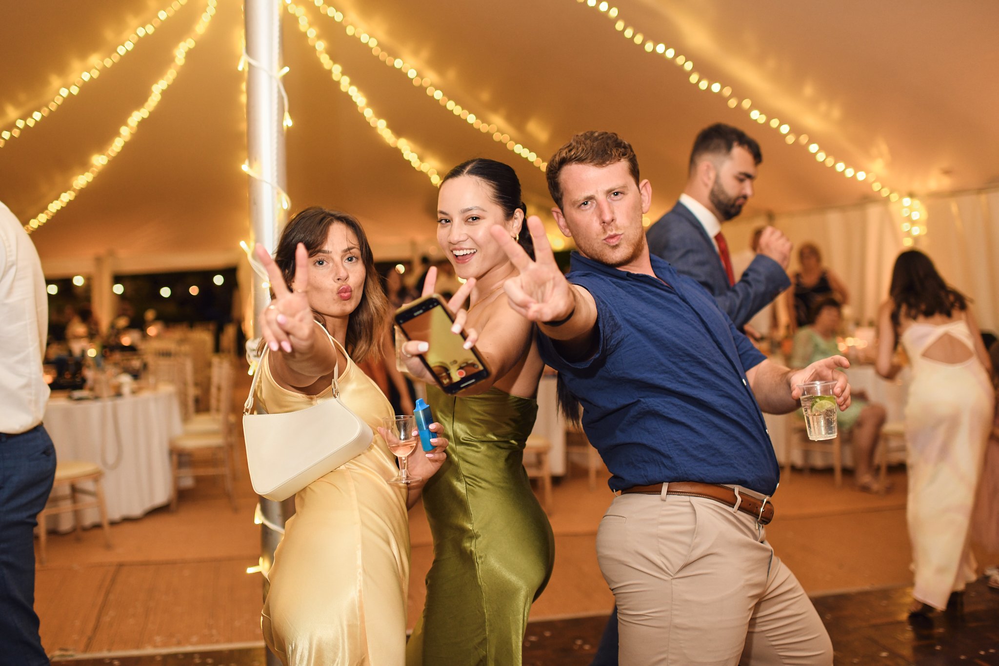 Three people dancing at a decorated party, two women and one man, all making peace signs and holding drinks, under string lights in a festive indoor setting.