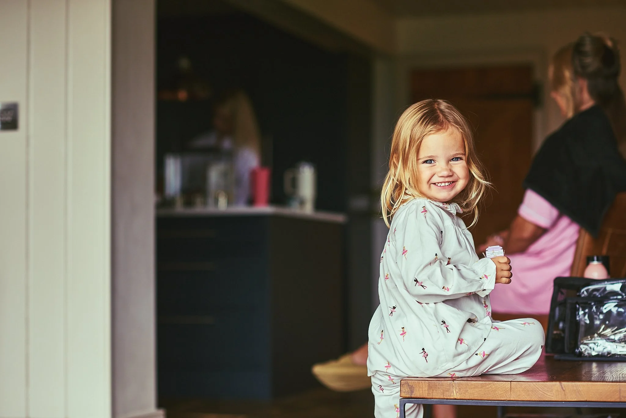 A young girl with blonde hair smiling at camera while sitting on a wooden table in a kitchen, with a woman in the background.