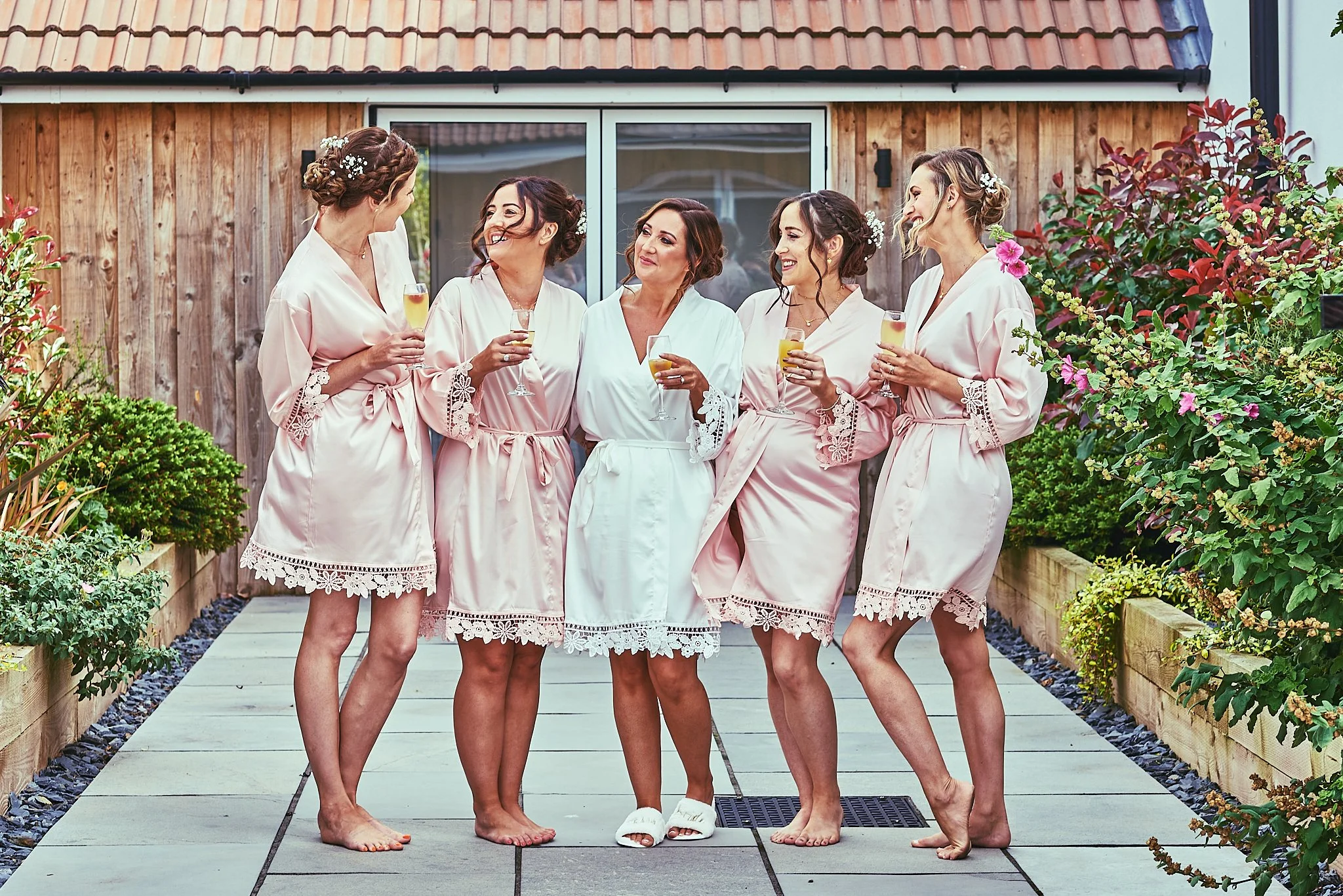 Five women in pink and white wedding robes, smiling and holding champagne glasses, standing outdoors on a paved walkway with plants on either side, in front of a wooden building with a sliding door.